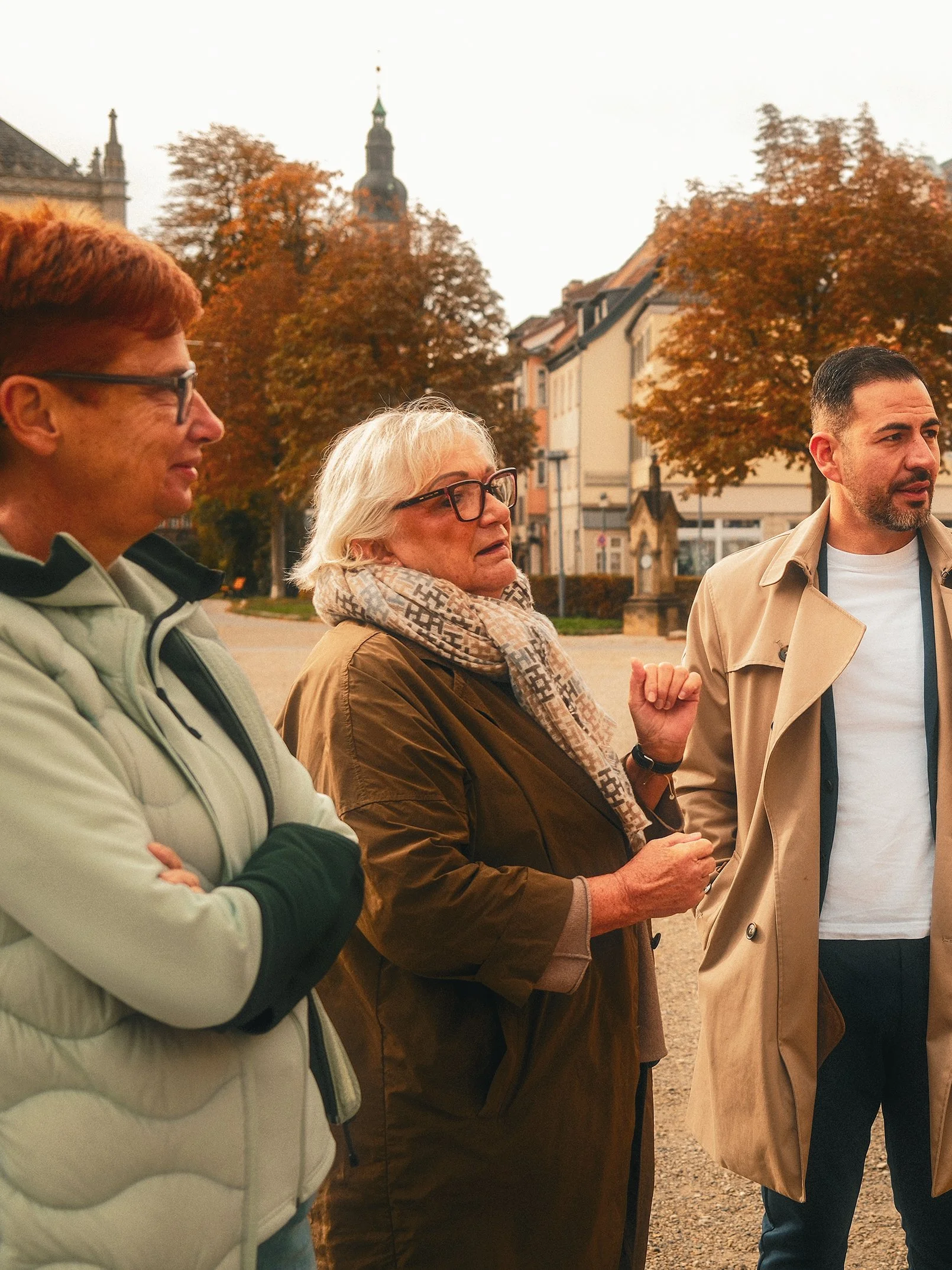 Drei Menschen, eine ältere Frau und zwei jüngere Männer, sprechen draußen vor historischen Gebäuden und Bäumen mit herbstlicher Laubfärbung.