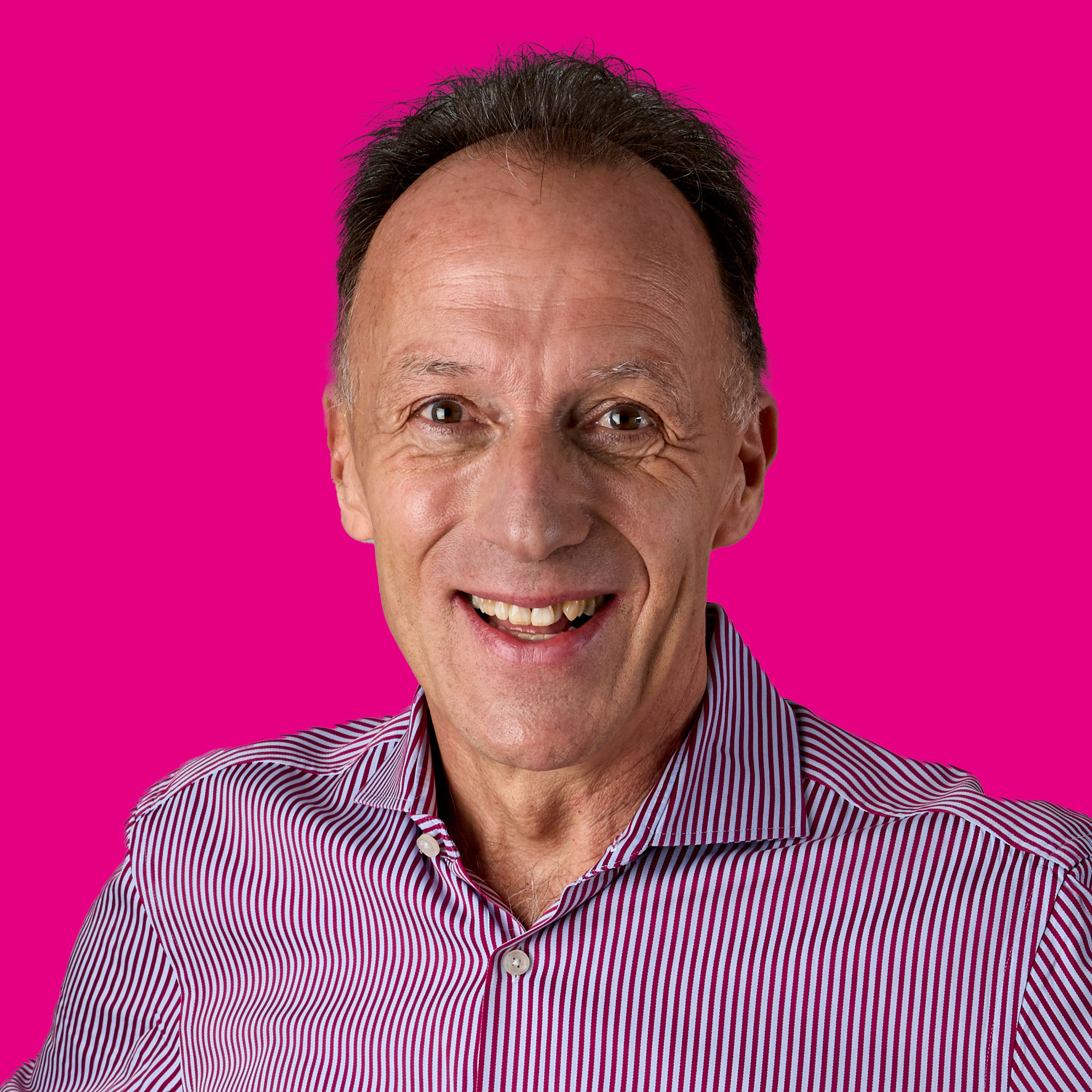 Ein älterer Mann lächelt freundlich, trägt ein rotes und weiß gestreiftes Hemd, vor pinkem Hintergrund.