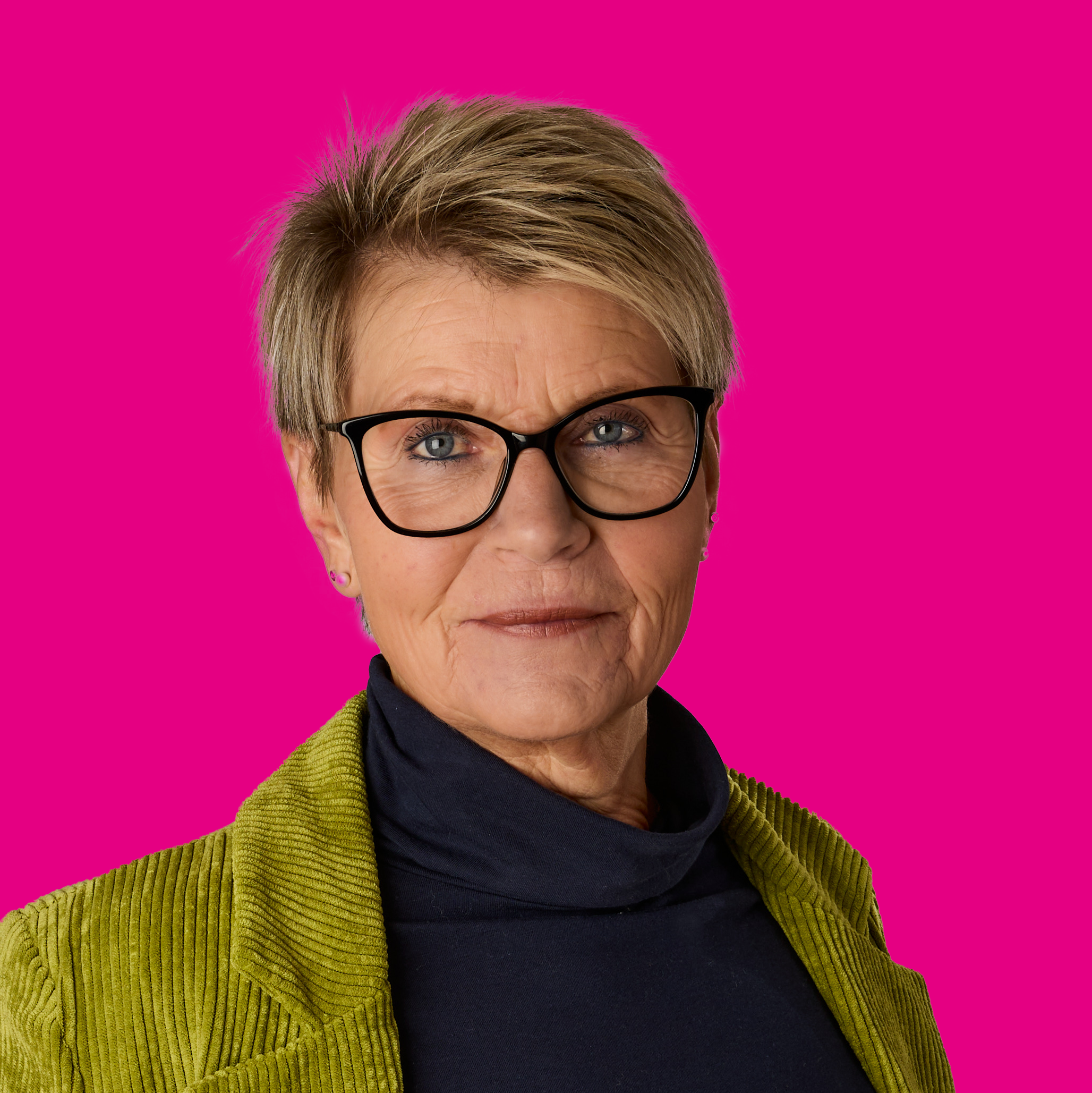 Porträt einer älteren Frau mit kurz blondem Haar, schwarzen Brillen, in einem dunkelblauen Tuch und grüner Jacke, vor pinkem Hintergrund.