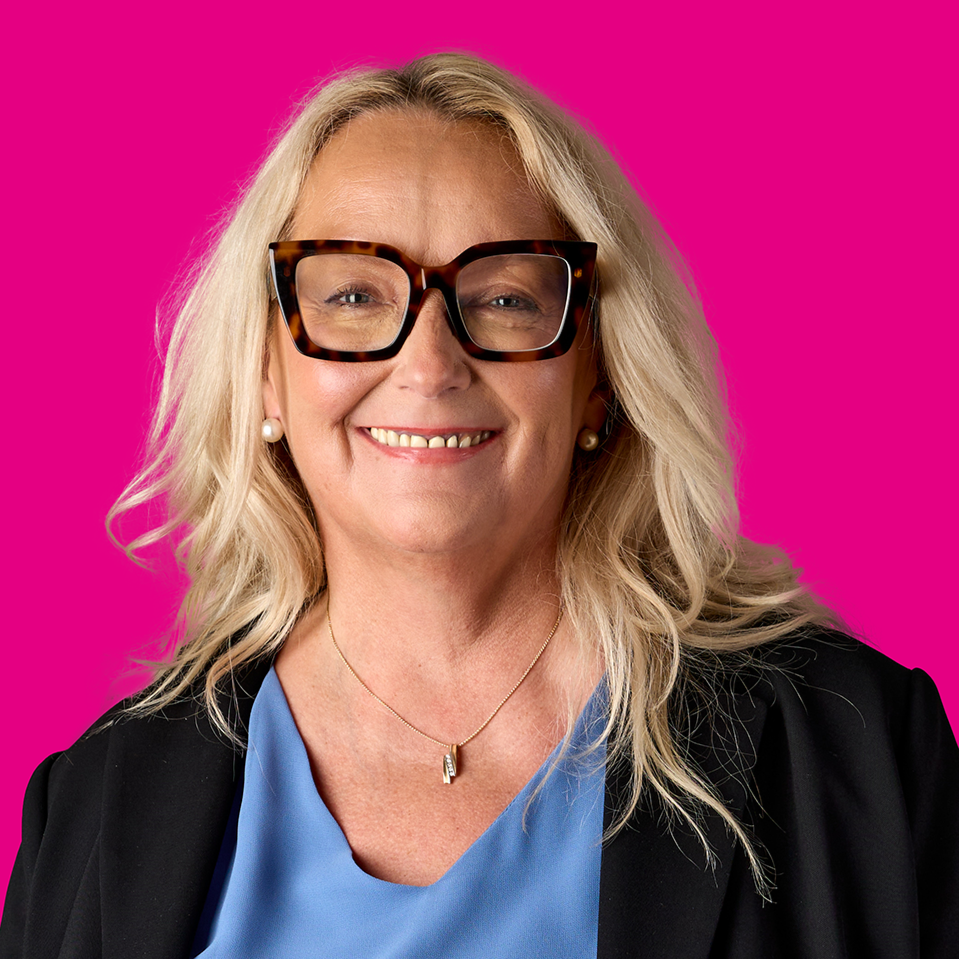 Eine lachende Frau mit langen blonden Haaren, großen braunen Brillen, Perlenohrringen, einem blauen Oberteil, einer schwarzen Jacke, einer goldenen Halskette und einem pinkfarbenen Hintergrund.