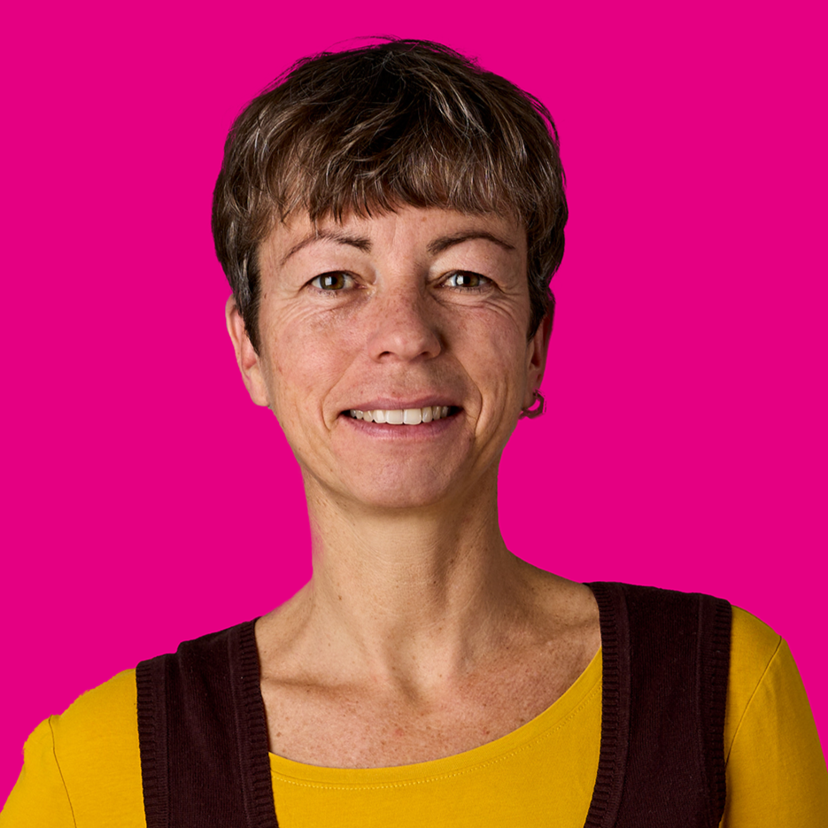 Eine lächelnde Frau mit kurzen braunen Haaren trägt ein gelbes Top und eine schwarze Weste vor einem pinken Hintergrund.