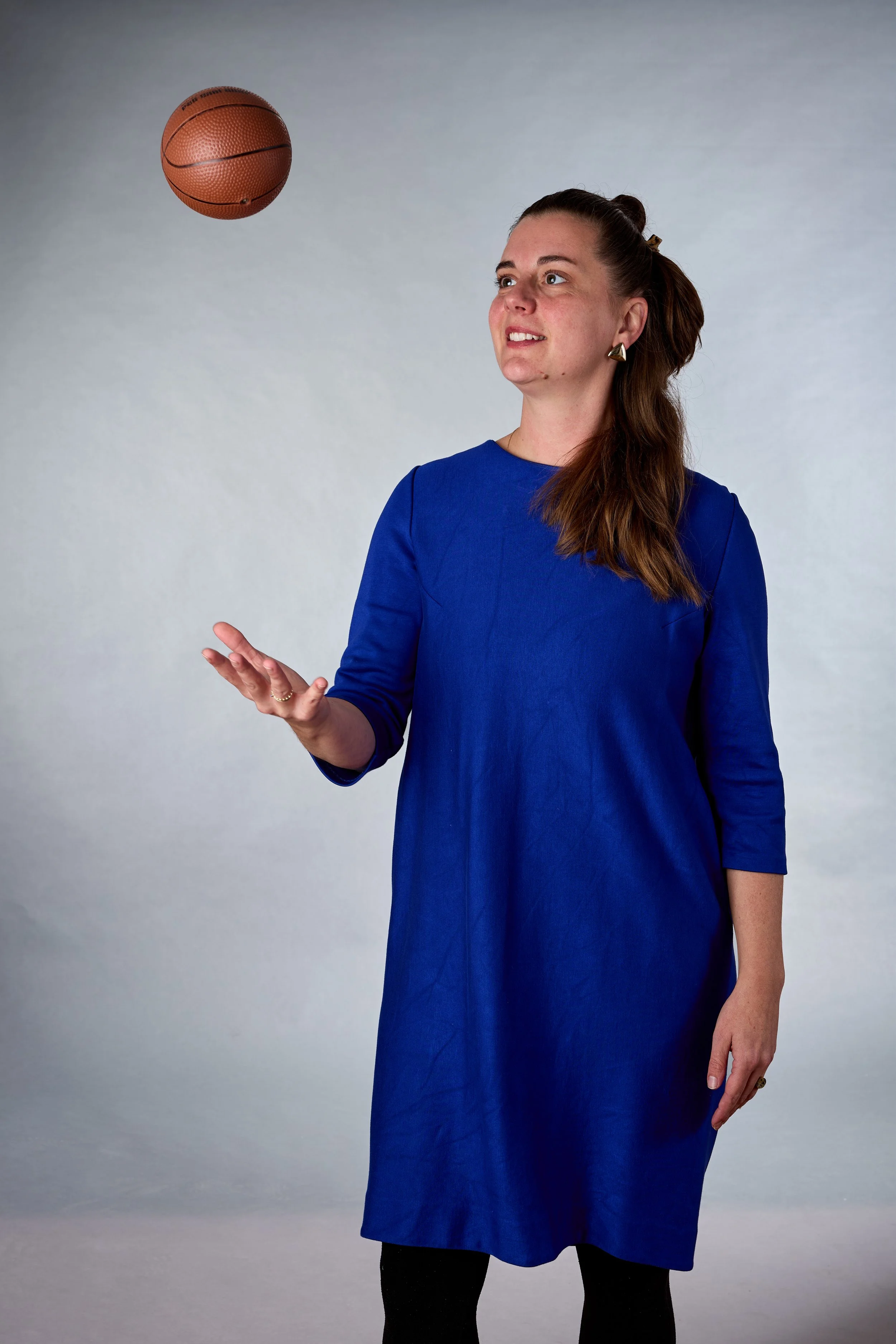 Eine Frau in einem blauen Kleid steht vor einem neutralen Hintergrund, während sie einen Basketball hält, der in der Luft schwebt.