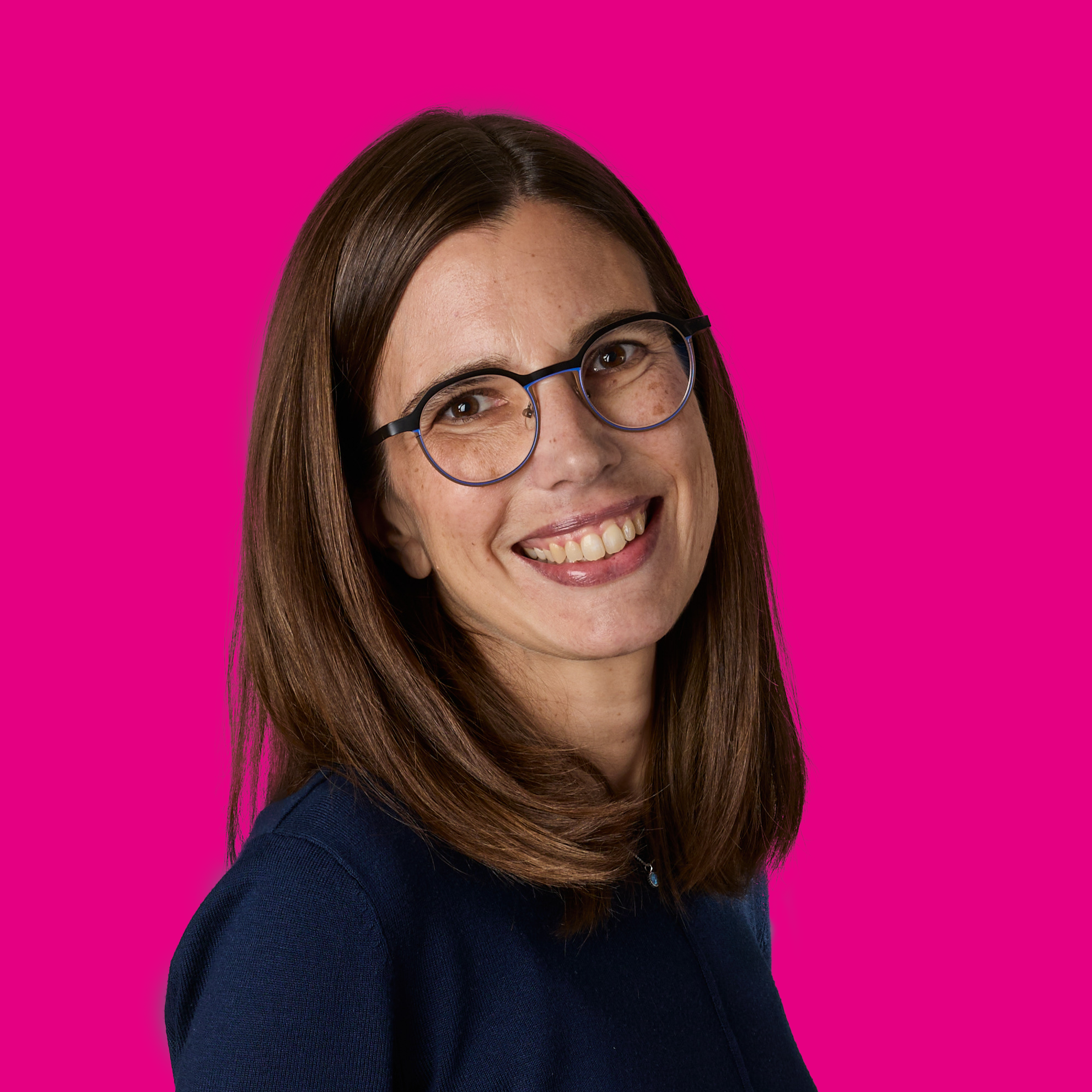 Freundliche junge Frau mit braunen Haaren, Brille und dunklem Top vor pinkem Hintergrund.