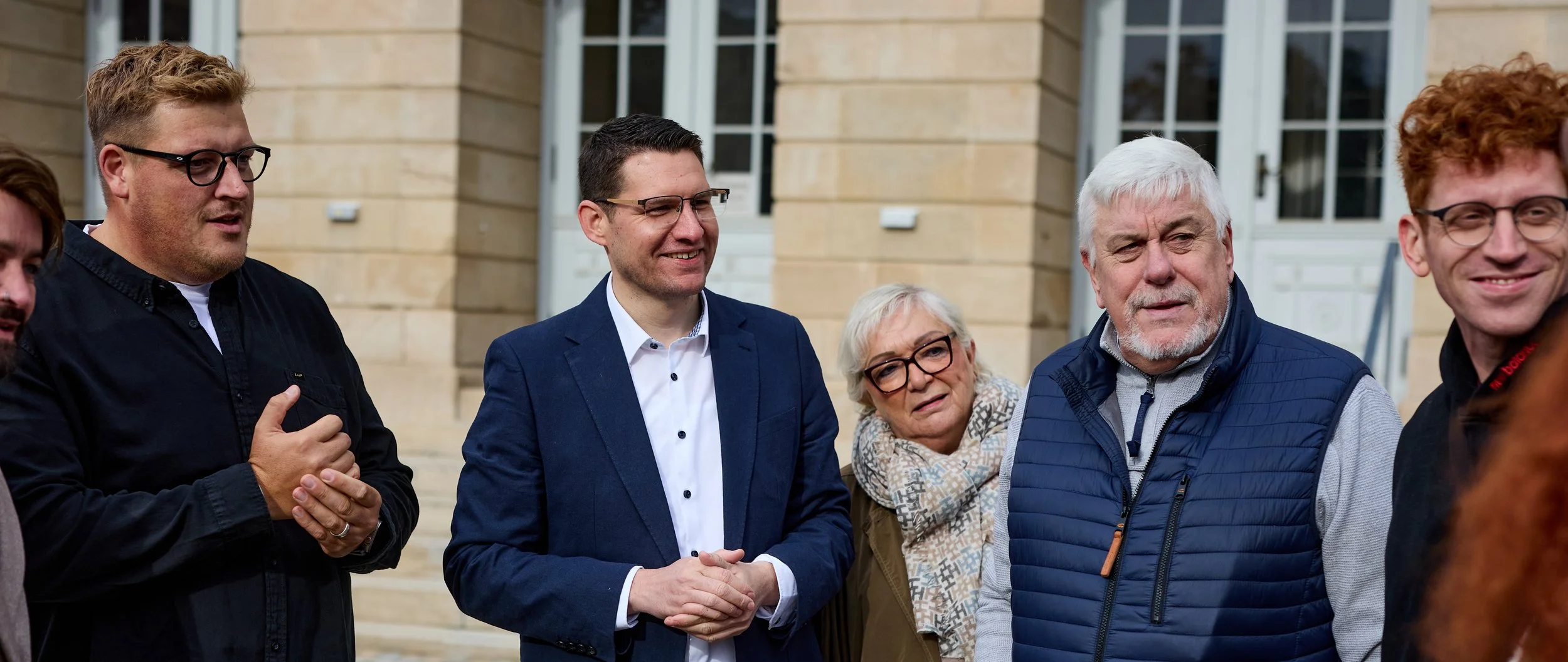Gruppe von fünf Menschen beim Gespräch vor einem Gebäude aus Sandstein, darunter zwei Männer mit Brillen, eine ältere Frau mit Brille und Schal, ein älterer Mann mit weißem Haar und einem blauen gesteppter Weste, alle sprechen oder lachen miteinander