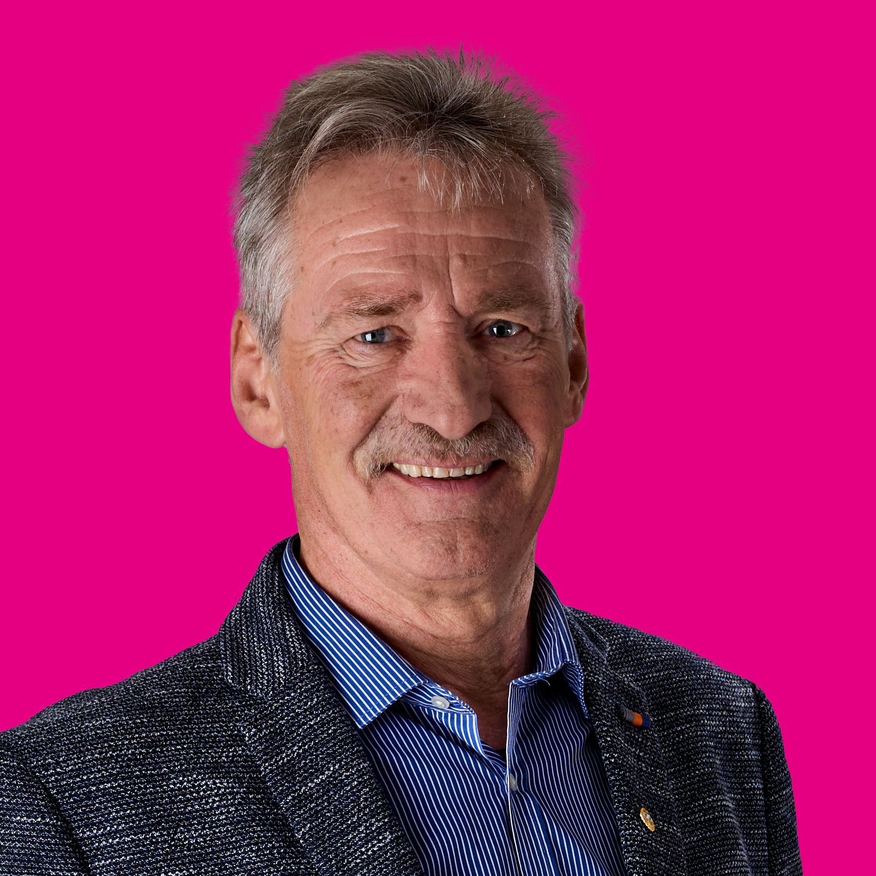 Porträt eines älteren Mannes mit grauem Haar, blauem Hemd und dunklem Sakko vor pinkem Hintergrund.