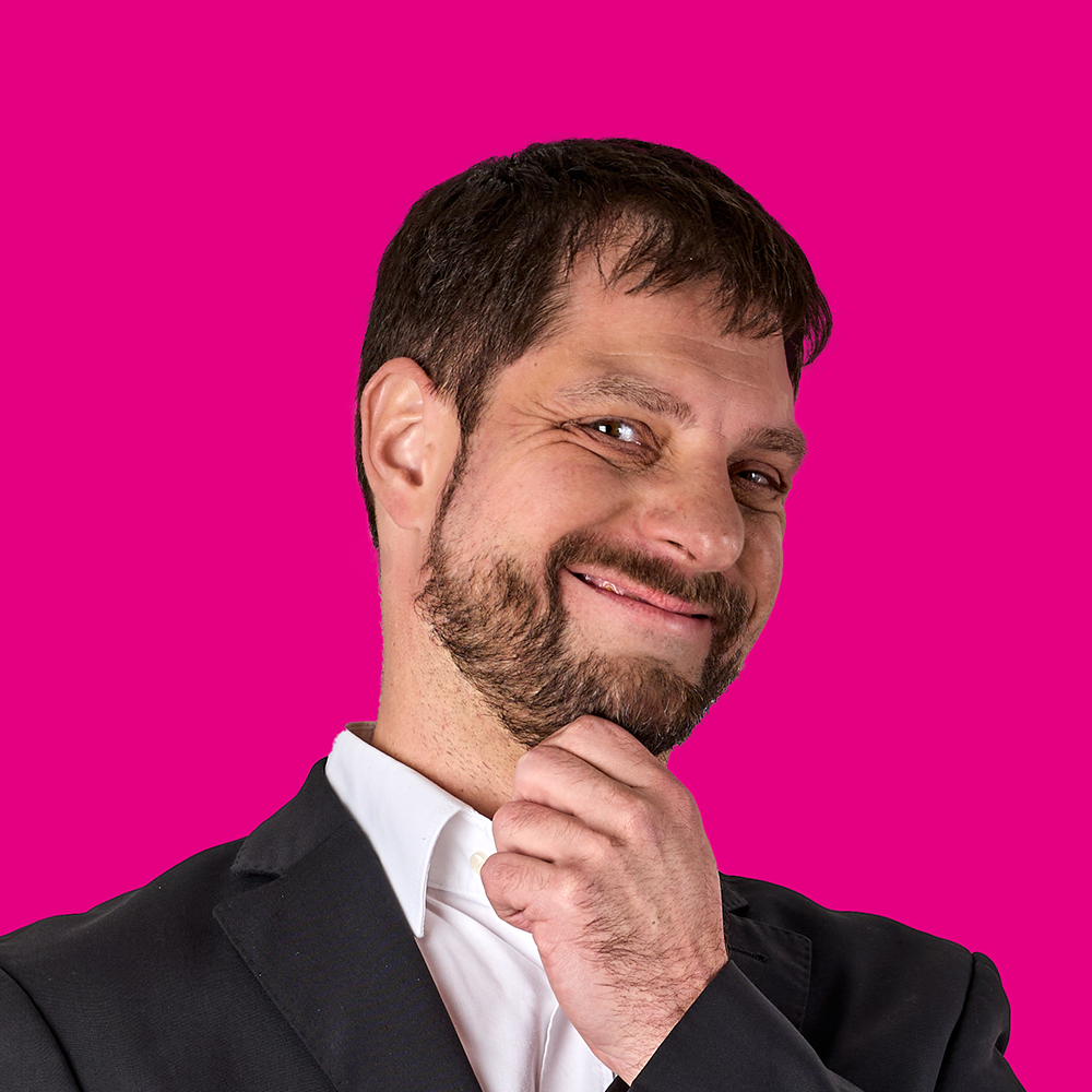 Ein lachender Mann mit dunklem Haar, Bart und Anzug vor pinkem Hintergrund, der nachdenklich wirkt.