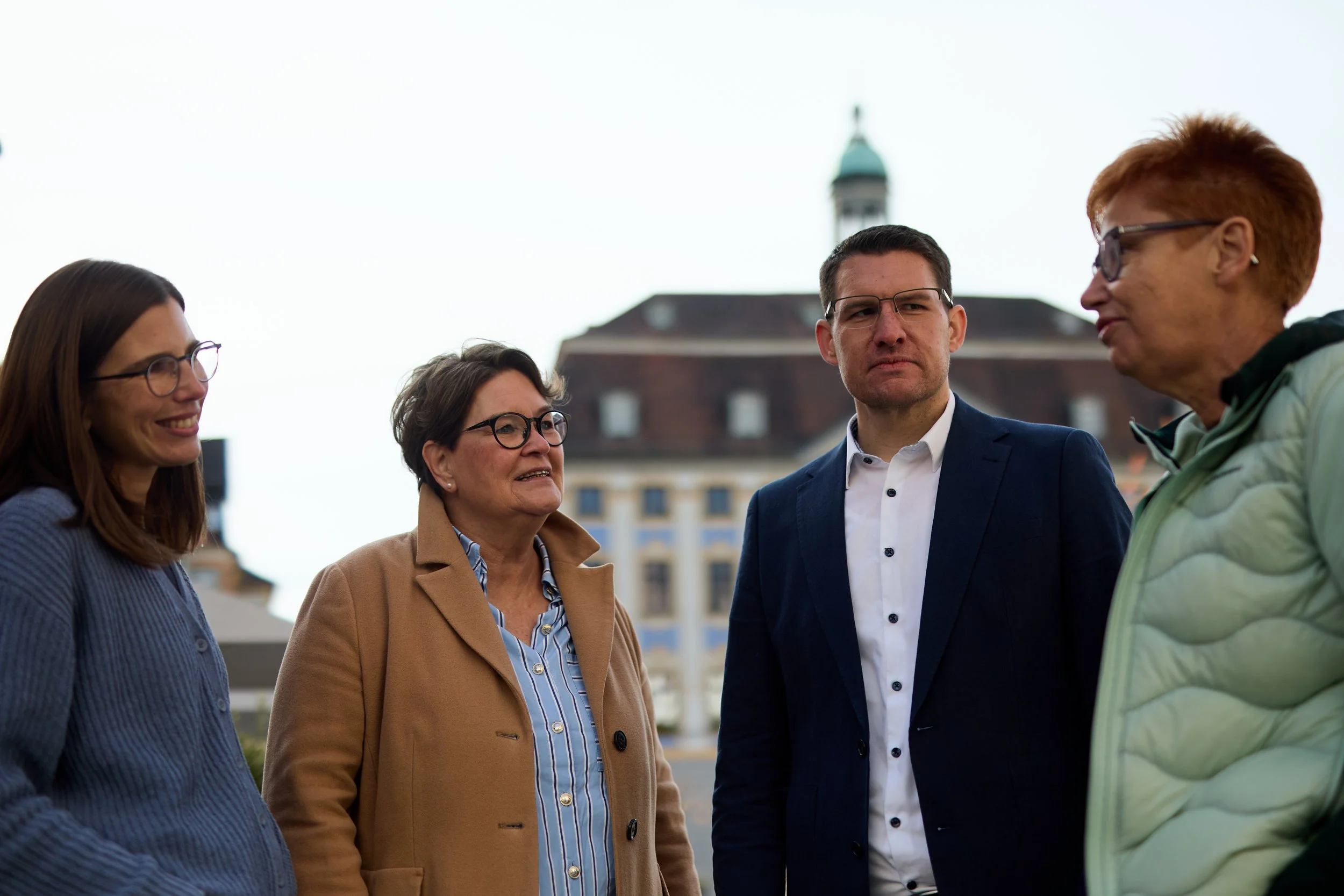 Fünf Menschen im Gespräch im Freien, im Hintergrund ein historisches Gebäude mit Uhrturm.