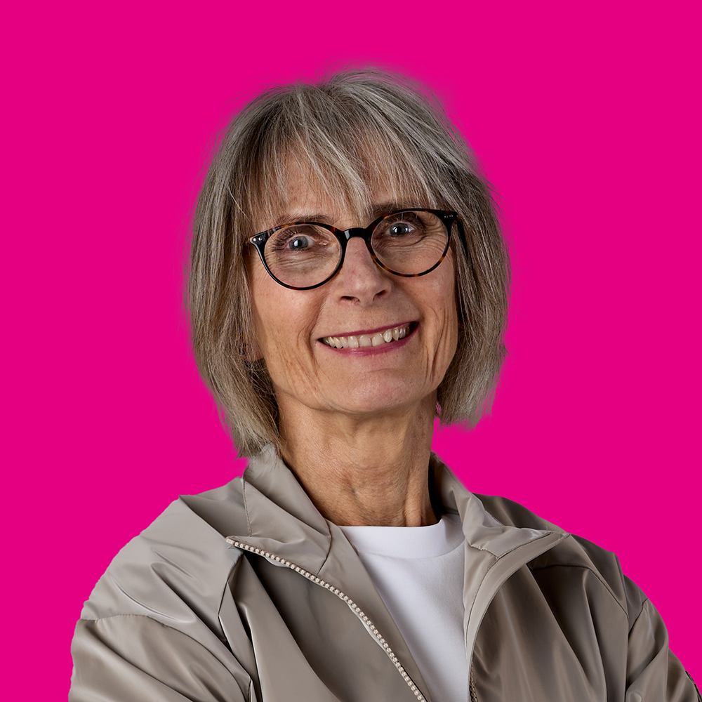 Porträt einer älteren Frau mit Brille, grauem Haar, lachend, vor pinkem Hintergrund.