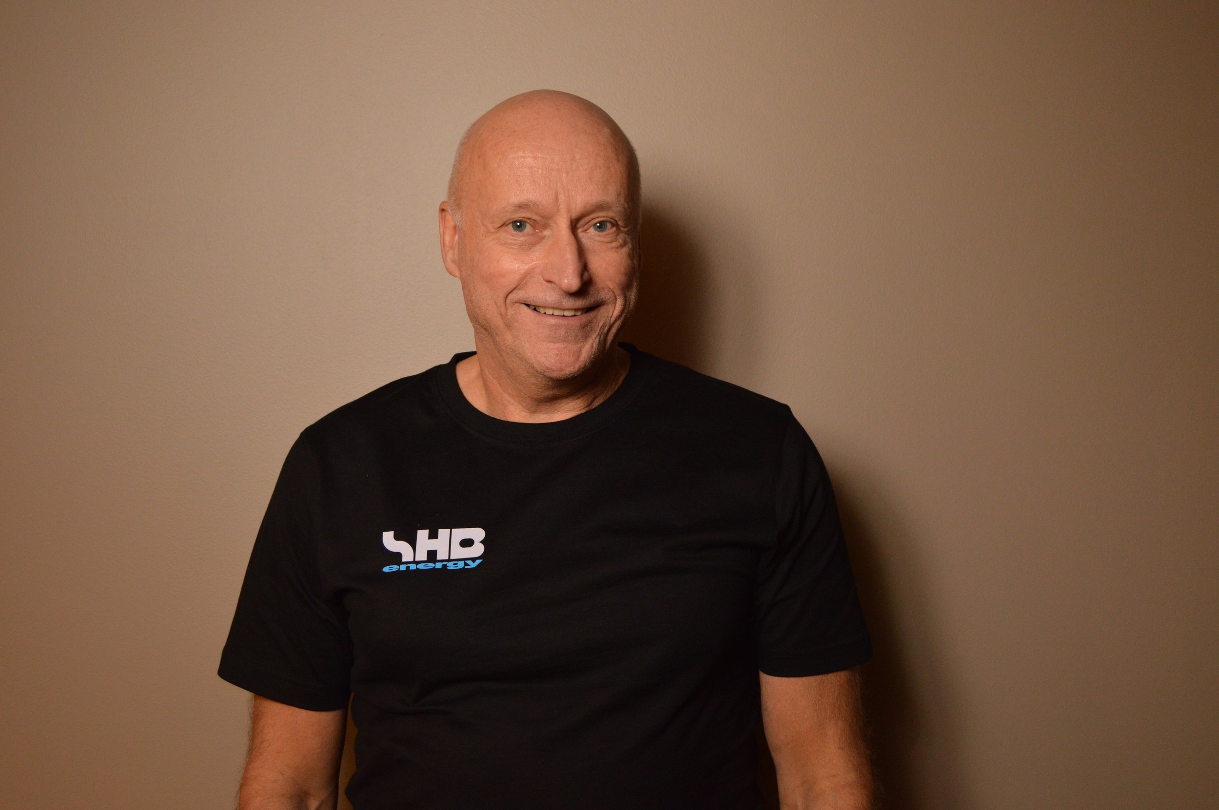 Smiling older man with a bald head wearing a black t-shirt with 'HB energy' logo, standing against a plain beige wall.
