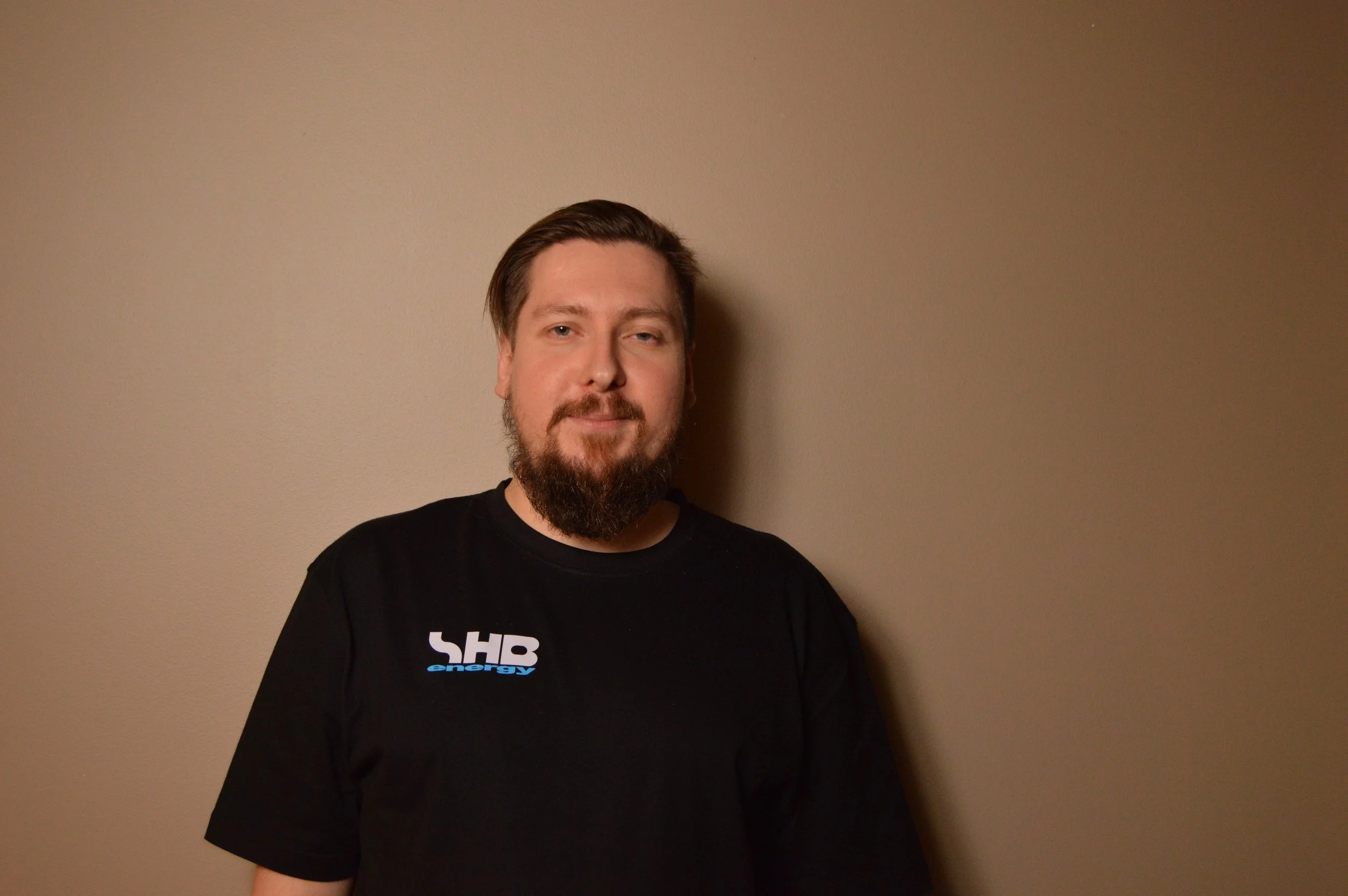 A man with a beard and mustache standing against a beige wall, wearing a black t-shirt with a logo that says 'SHB energy'.