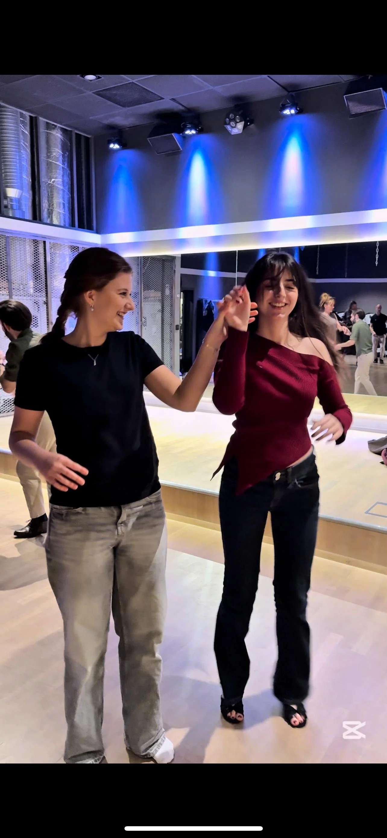 Two women dancing salsa and smiling in a dance studio with blue stage lights overhead, mirrors on the wall, and other people dancing in the background.