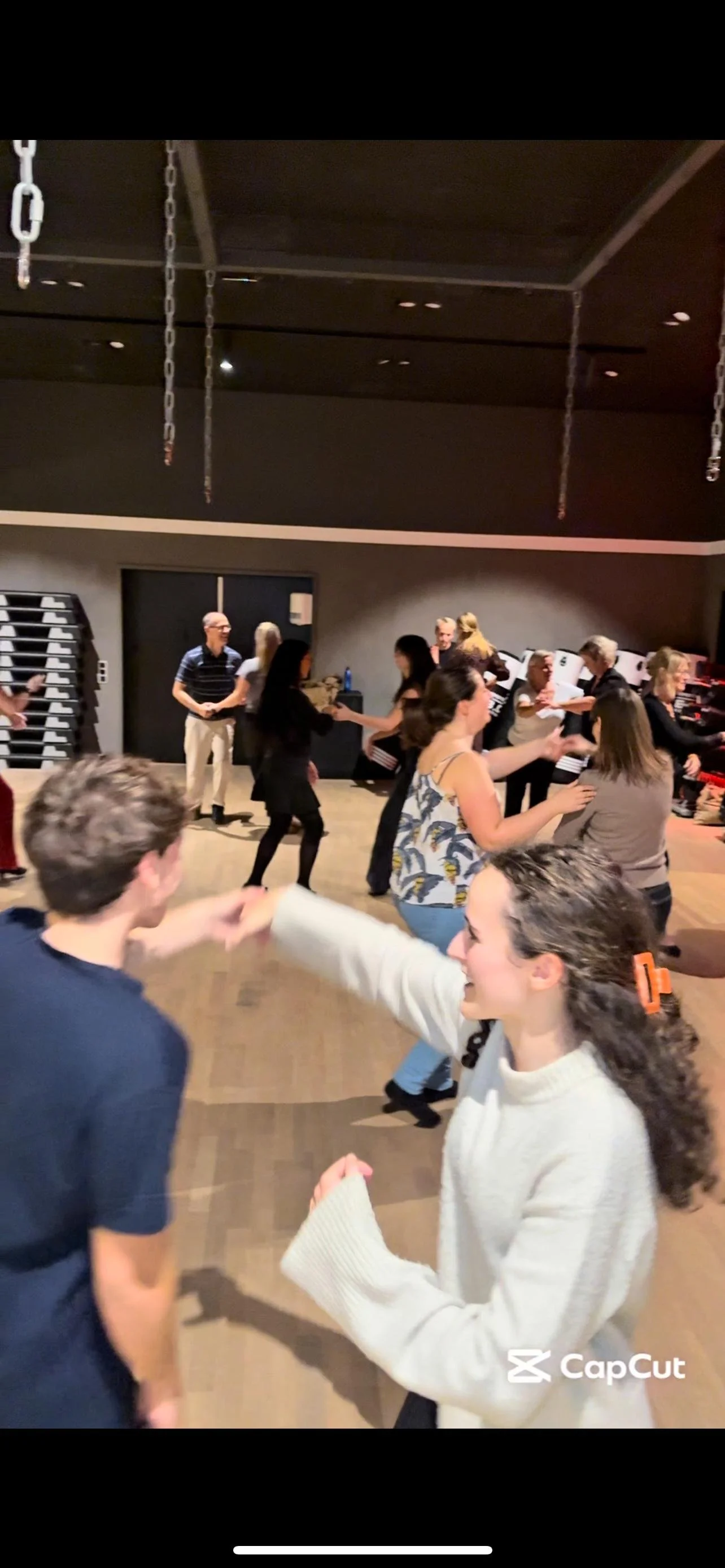 People dancing and socializing in a dimly lit indoor venue with a wooden floor and ceiling, some engaged in conversation or handshakes, creating a lively and festive atmosphere.