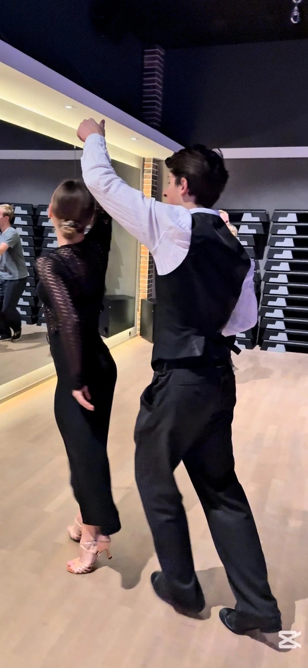 A man and a woman dancing on a wooden floor in a dimly lit room, with the man facing towards the woman and raising his arm, while the woman has her back to the camera and is wearing a black, sheer, long-sleeved dress with high heels.