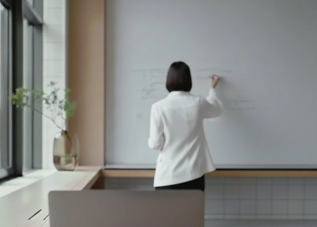 A woman in a white blazer writing on a whiteboard in a bright, modern classroom or office.