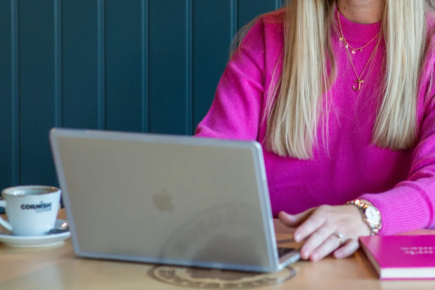 A woman in a bright pink sweater is sitting at a wooden table working on a silver laptop. She is wearing gold jewelry, including a necklace with letters and a cross, as well as a gold watch and rings. There is a white coffee cup with