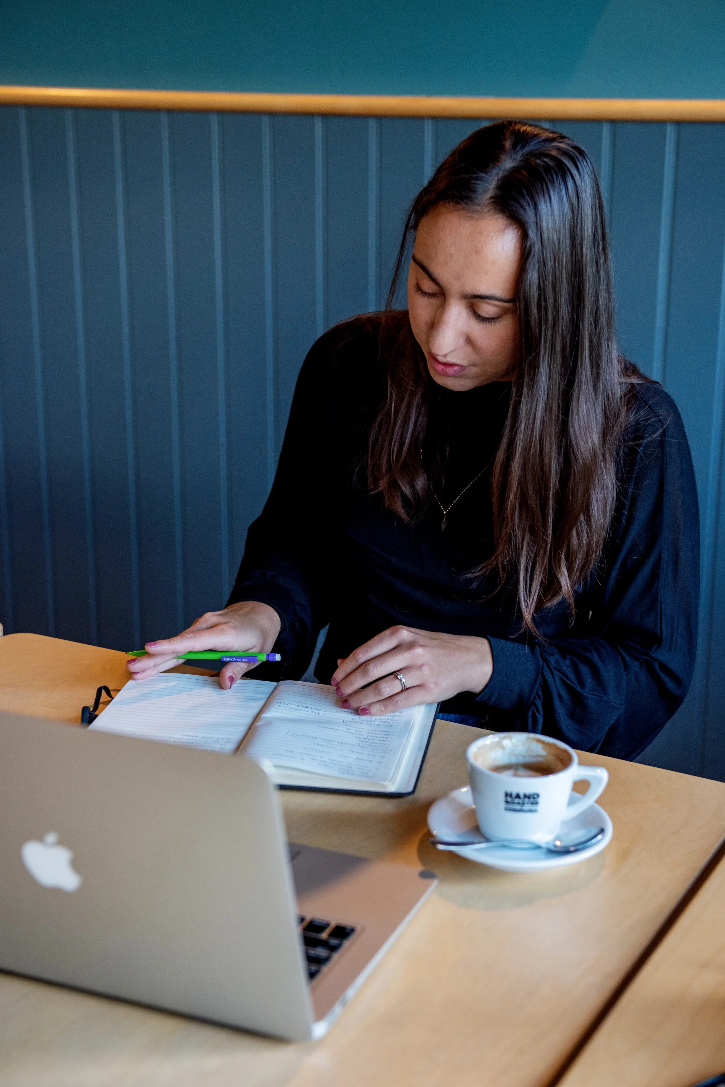 A woman with long brown hair, wearing a black shirt, sitting at a wooden table in a cafe, reading a notebook with a pen in her hand. There is an open silver MacBook laptop and a white cup of coffee on a saucer on the table.