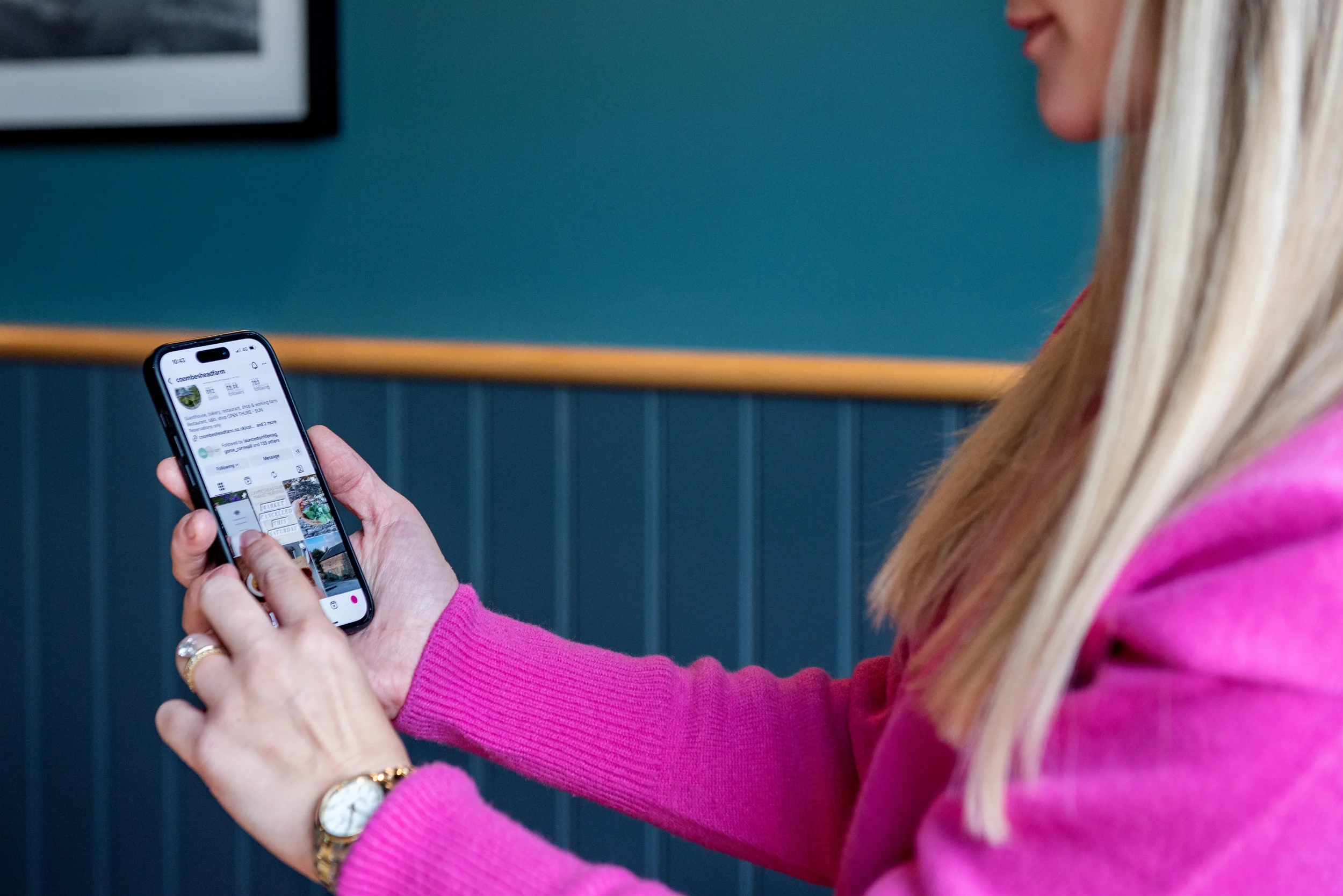 A woman with long blonde hair wearing a pink sweater using a smartphone to browse Instagram in a room with teal walls and wood paneling.