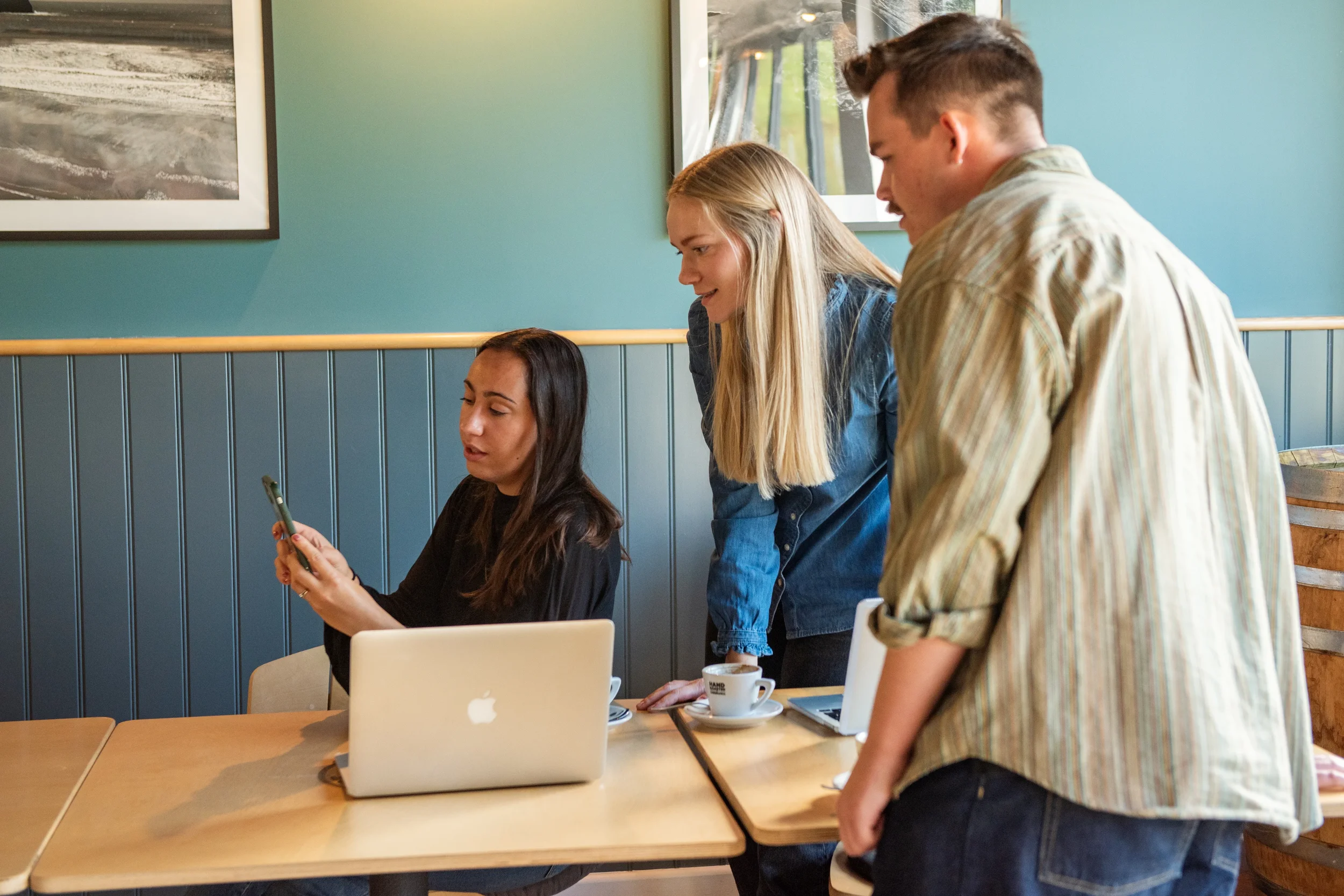 Three young adults, two women and one man, are gathered around a table in a café discussing something on a phone the woman seated is holding. The woman seated has long dark hair and is wearing a black top, while the woman standing has long blonde hair and wears a denim shirt. The man standing has short hair and is wearing a striped shirt. There are coffee cups, an open laptop, and a notebook on the table.