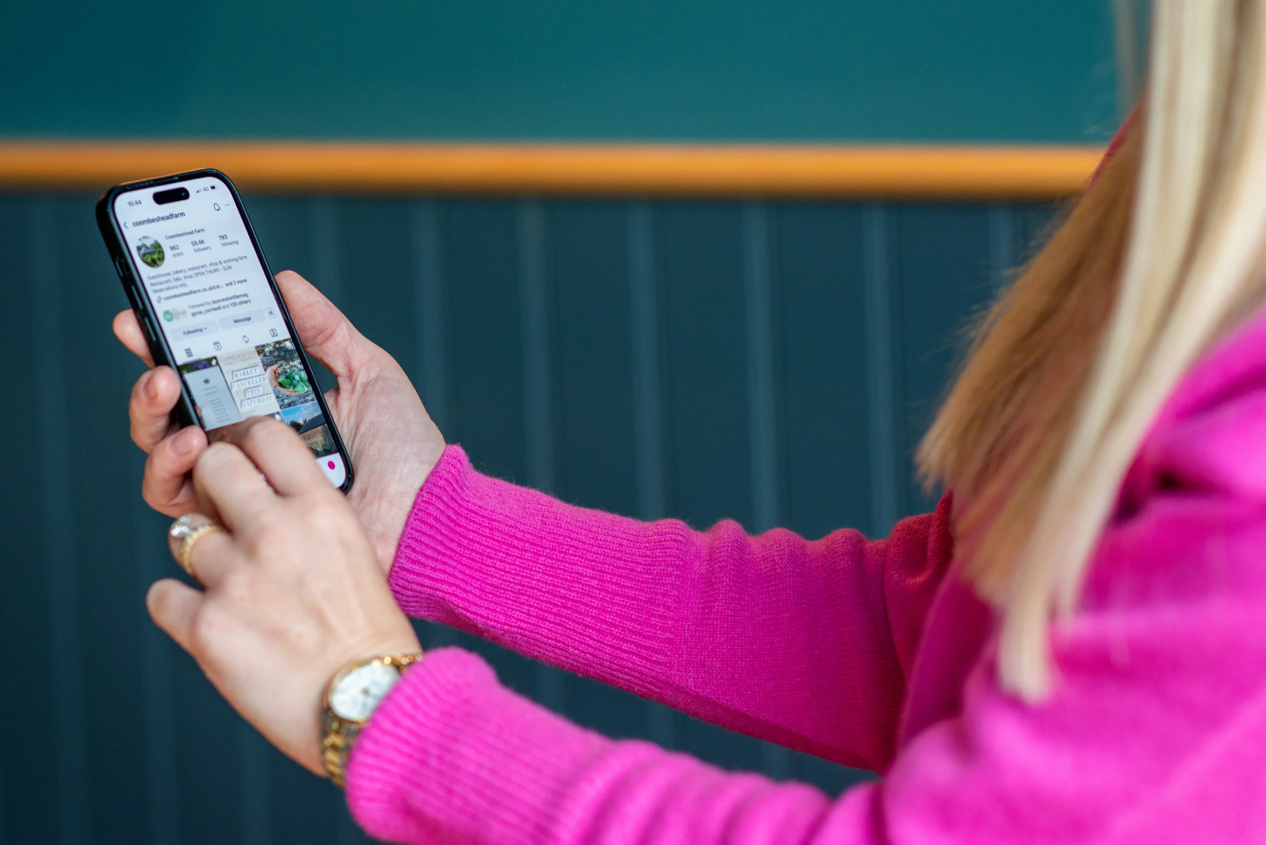 Person in pink sweater holding a smartphone, browsing a social media profile.