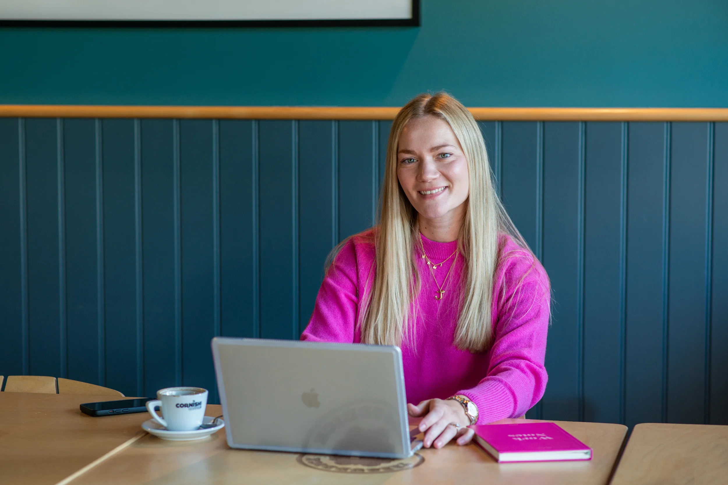 A woman with long blonde hair sitting at a wooden table, working on a silver laptop. She is wearing a bright pink sweater, layered necklaces, a gold watch, and has a coffee cup, a pink notebook, and a phone on the table. The background features a teal wall with wooden wainscoting.