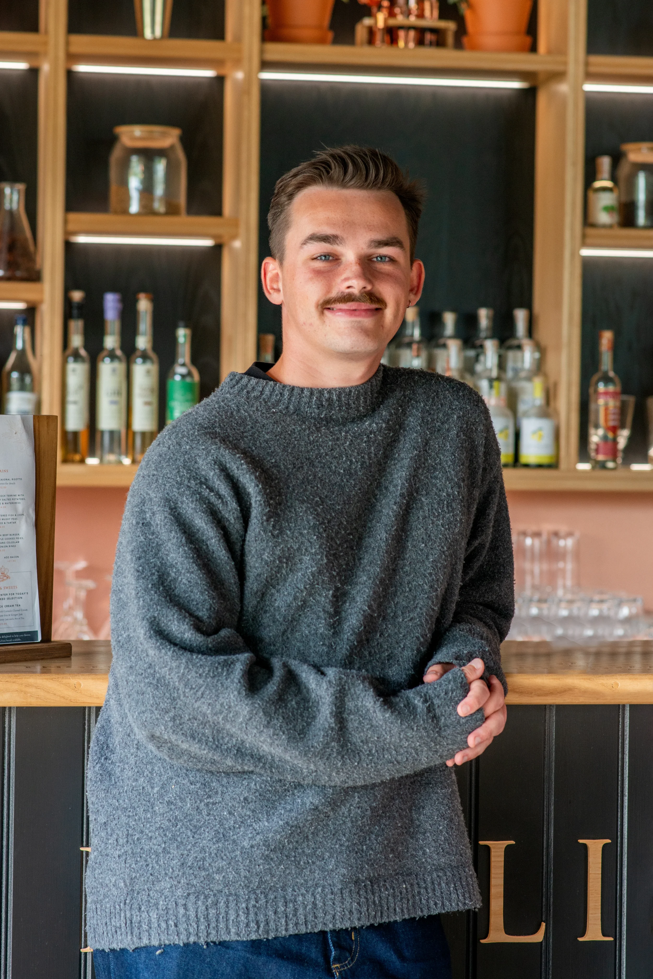 A young man with a mustache and short brown hair, wearing a dark gray sweater, standing in front of a bar with bottles of liquor on shelves behind him.