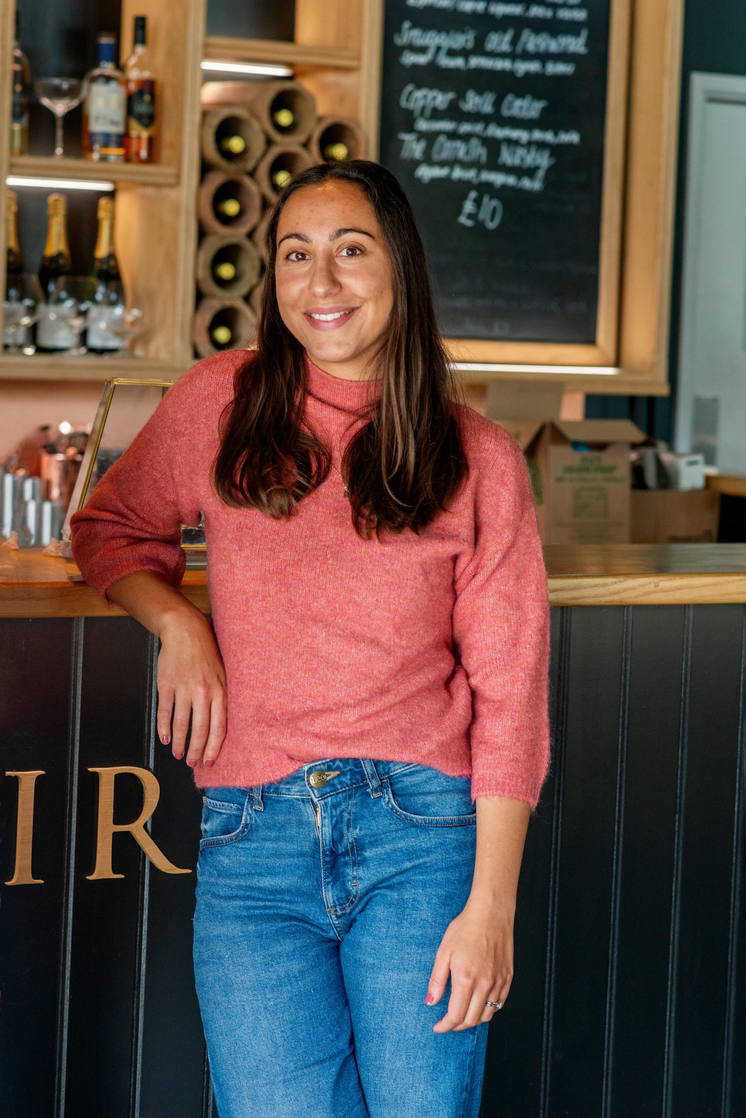 A woman with long dark hair wearing a pink sweater and blue jeans, standing inside a bar or cafe, smiling at the camera.