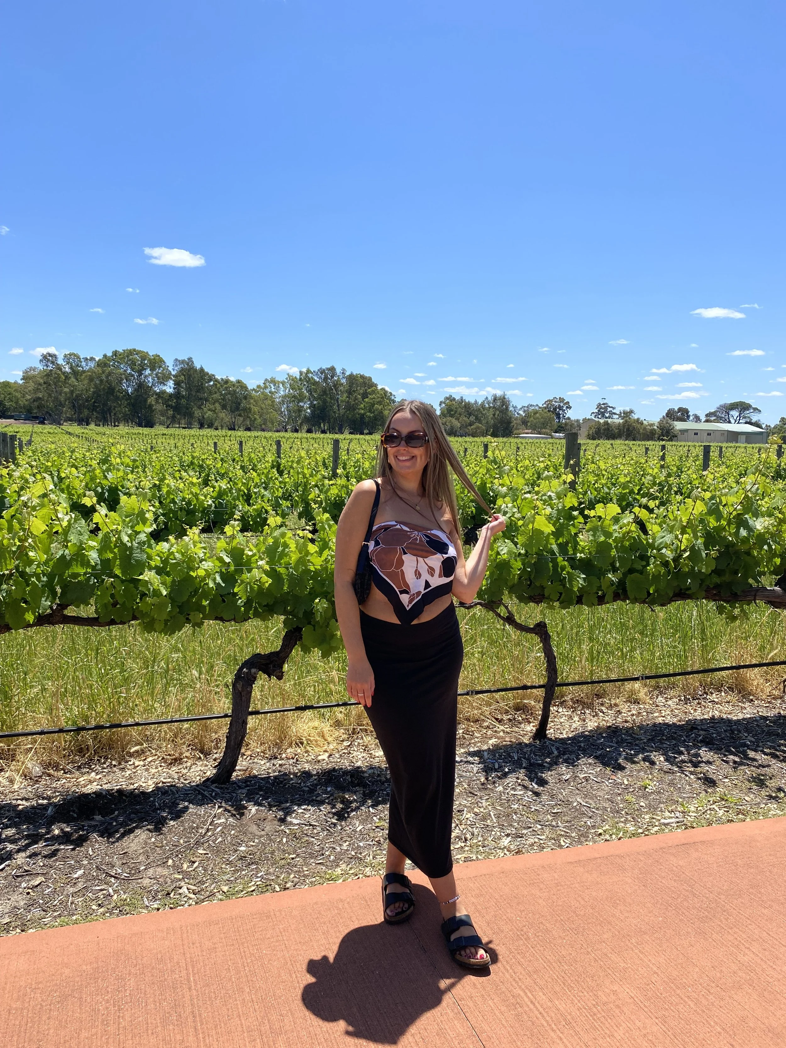 A woman wearing sunglasses, a black skirt, and a sleeveless top with a geometric pattern stands on a sidewalk in a vineyard under a bright blue sky with scattered clouds, smiling and holding her hair.