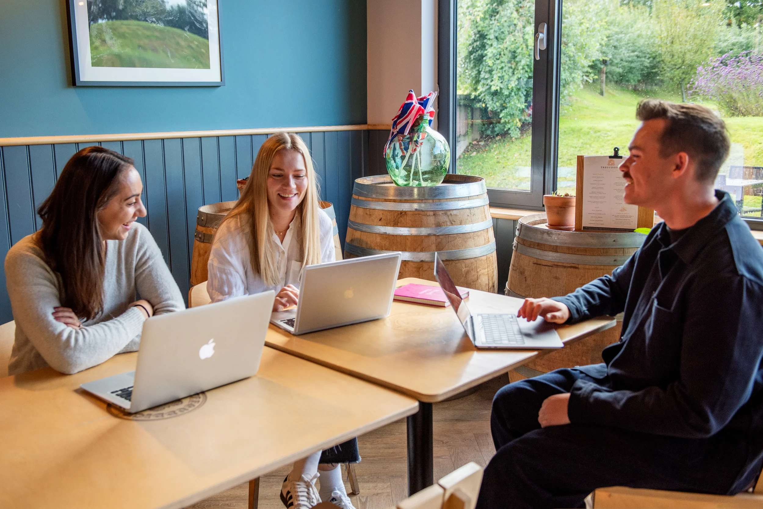 Three young adults sitting around a table in a room with large windows, laughing and working on laptops.