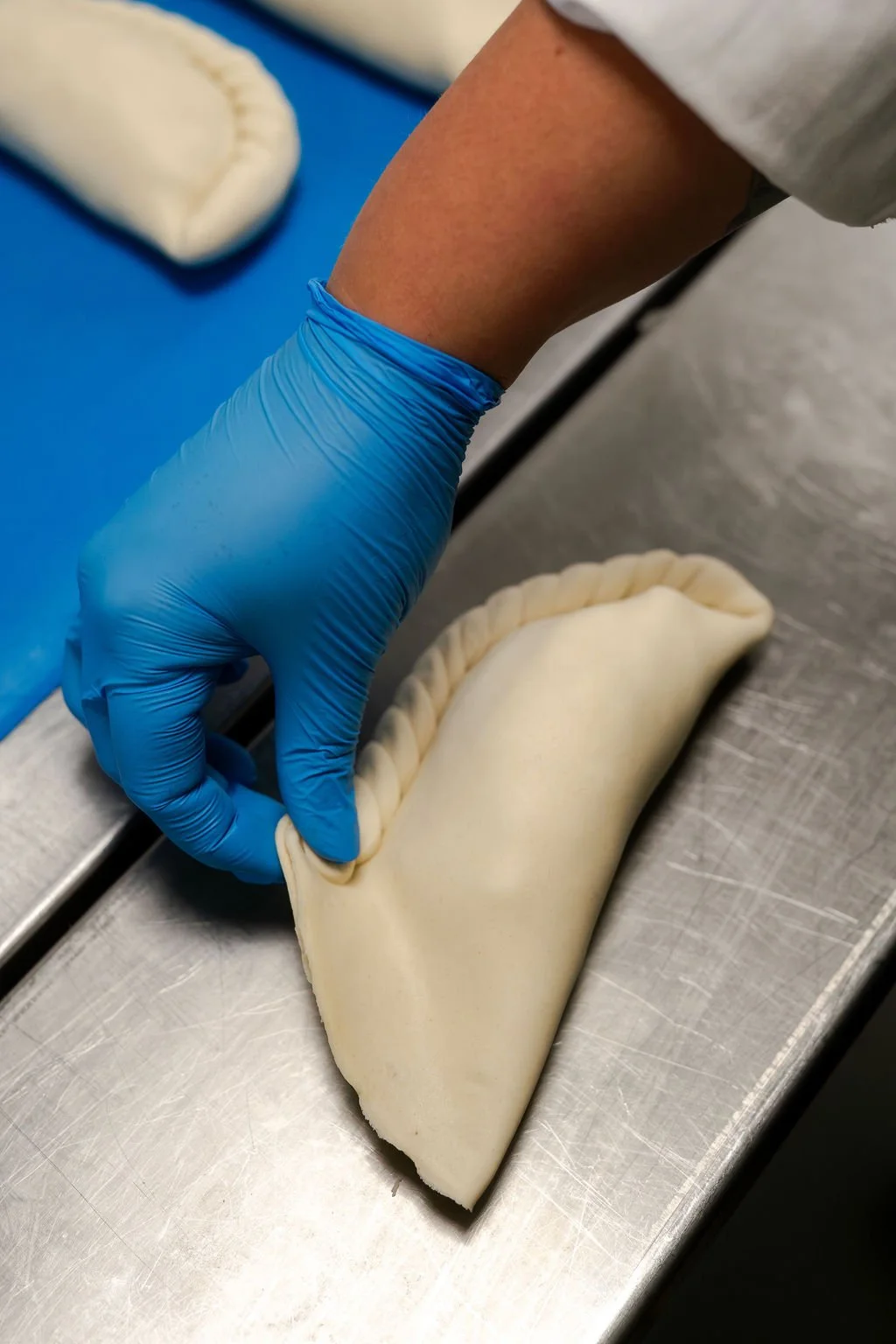 Person wearing blue gloves folding raw pie or pastry dough on a metal surface.