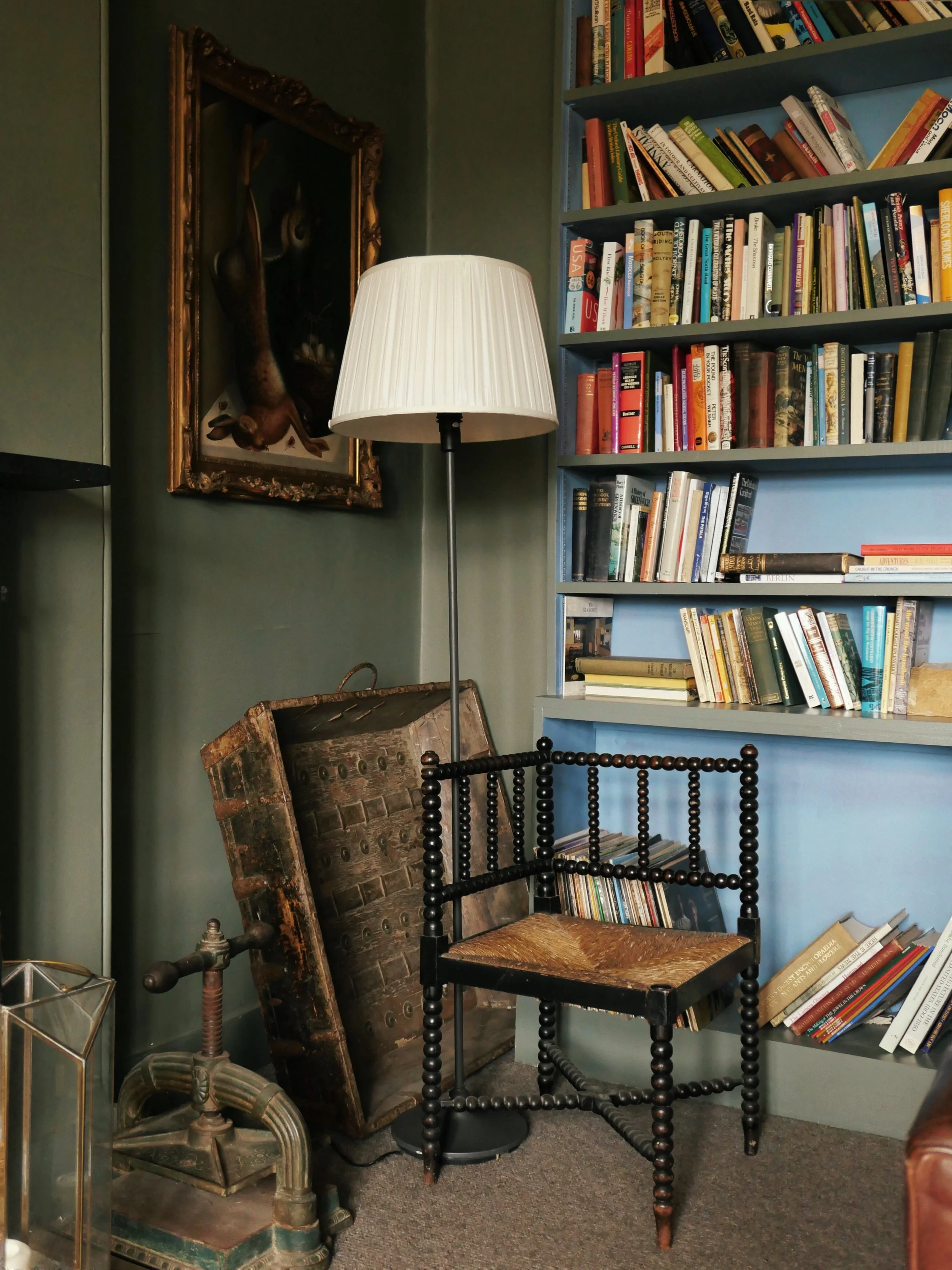 An interior scene with a bookshelf filled with books on the right side, a floor lamp with a white shade in the center, an ornate picture frame with a painting or photograph, an antique wooden trunk leaning against the wall, and an old fashioned coin bank or weight on the floor in the lower left corner.