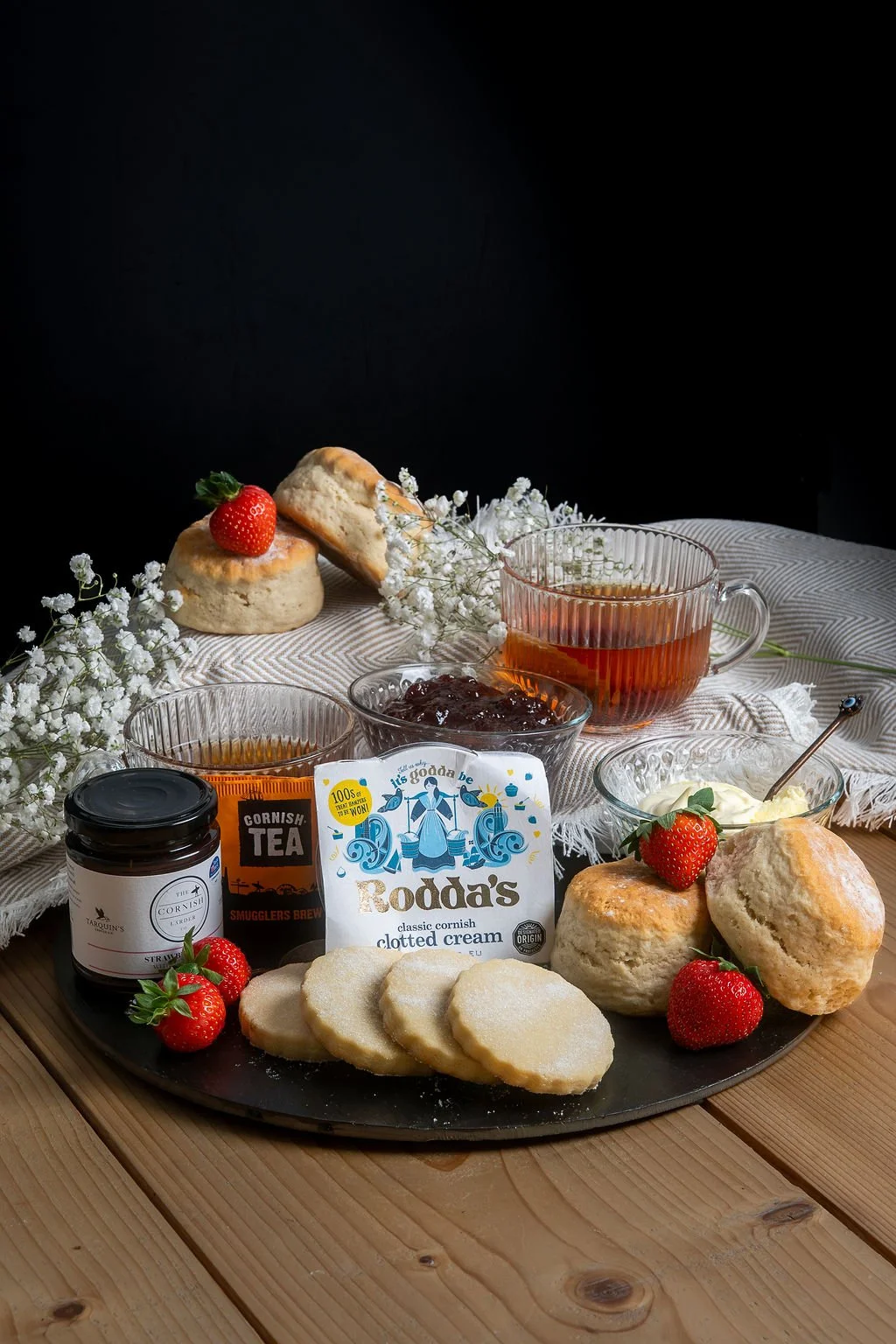 A breakfast spread on a black tray includes scones topped with strawberries, a jar of strawberry jam, a bowl of clotted cream, cookies, two glasses of tea, and a bowl of preserves. The setup is decorated with white flowers on a wooden table with a white cloth and a dark background.