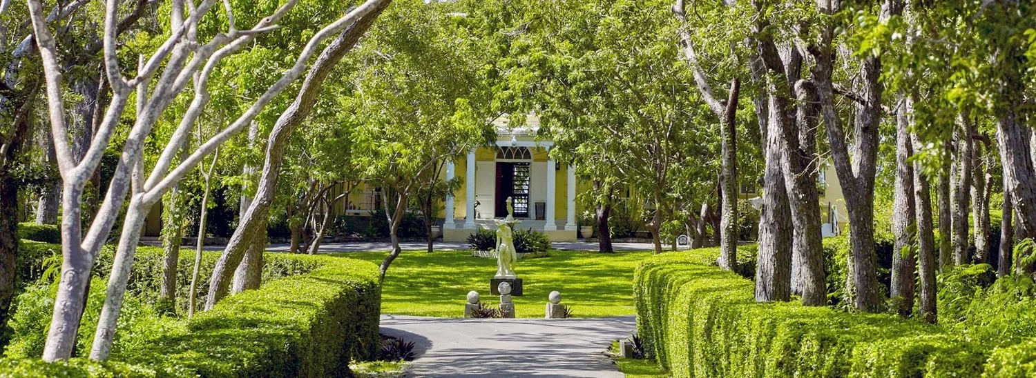 Garden pathway leading to a white house surrounded by trees and well-manicured bushes, with a statue in the center.