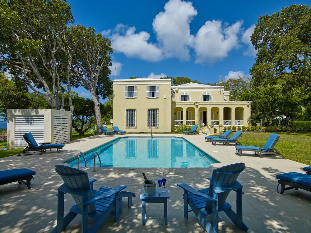 A large swimming pool surrounded by blue lounge chairs in a yard with green grass and large trees, with a two-story yellow house with white trim in the background under a partly cloudy sky.
