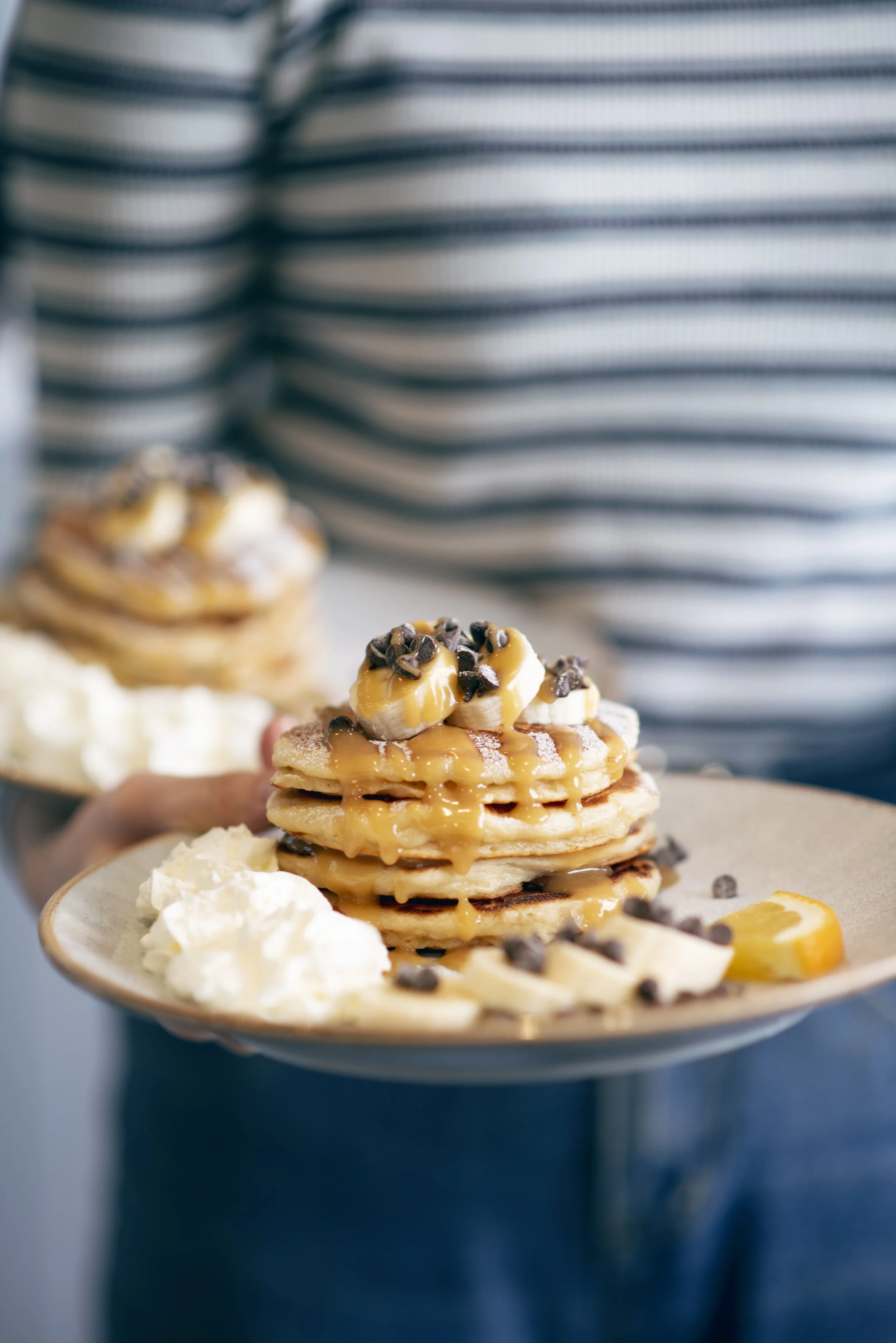 A person holding a plate with a stack of four golden brown pancakes topped with sliced bananas, chocolate chips, and caramel drizzle. There is whipped cream on the side of the plate and another similar stack in the background.