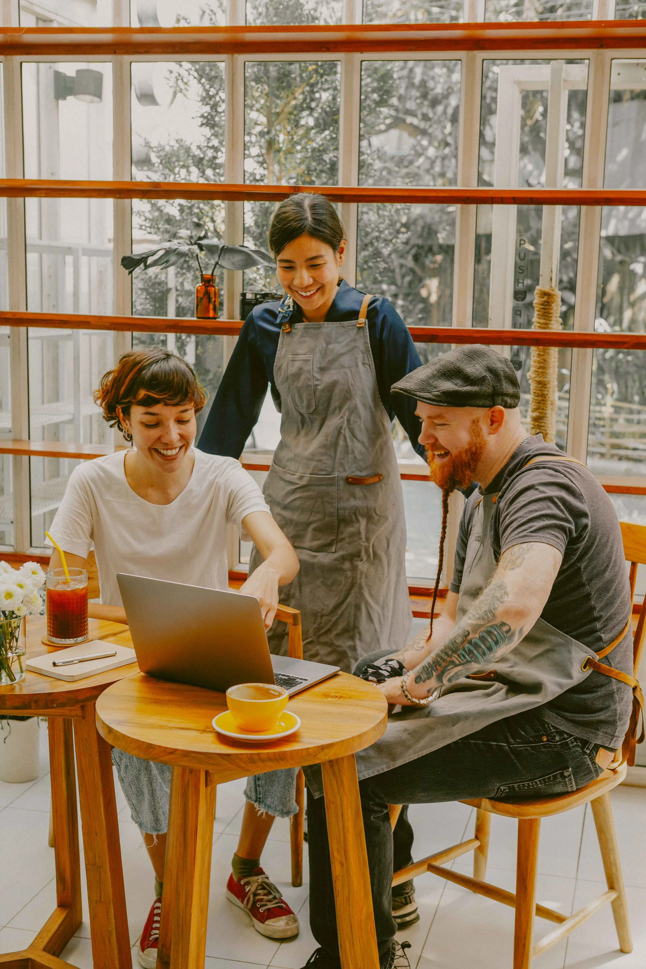 Tres personas en una cafetería mirando una computadora portátil y sonriendo, mientras una camarera observa.