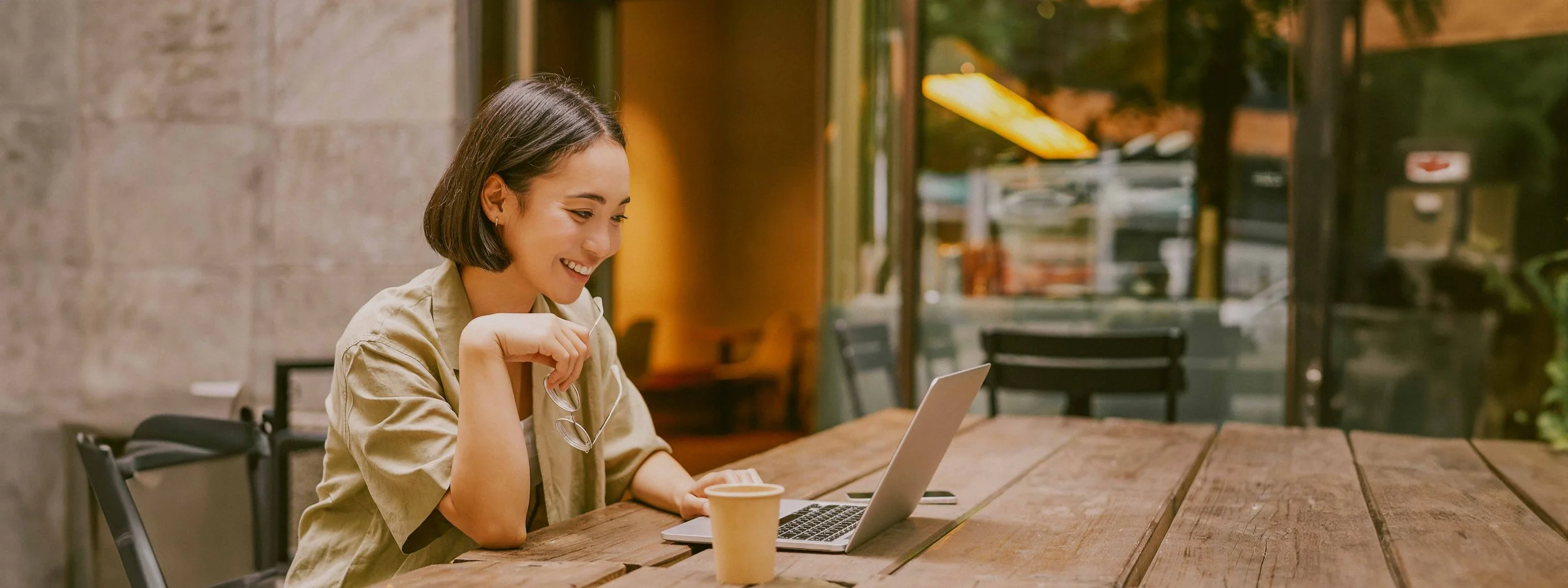 Mujer sonriendo usando una laptop en una cafetería, con una taza de café en la mesa.