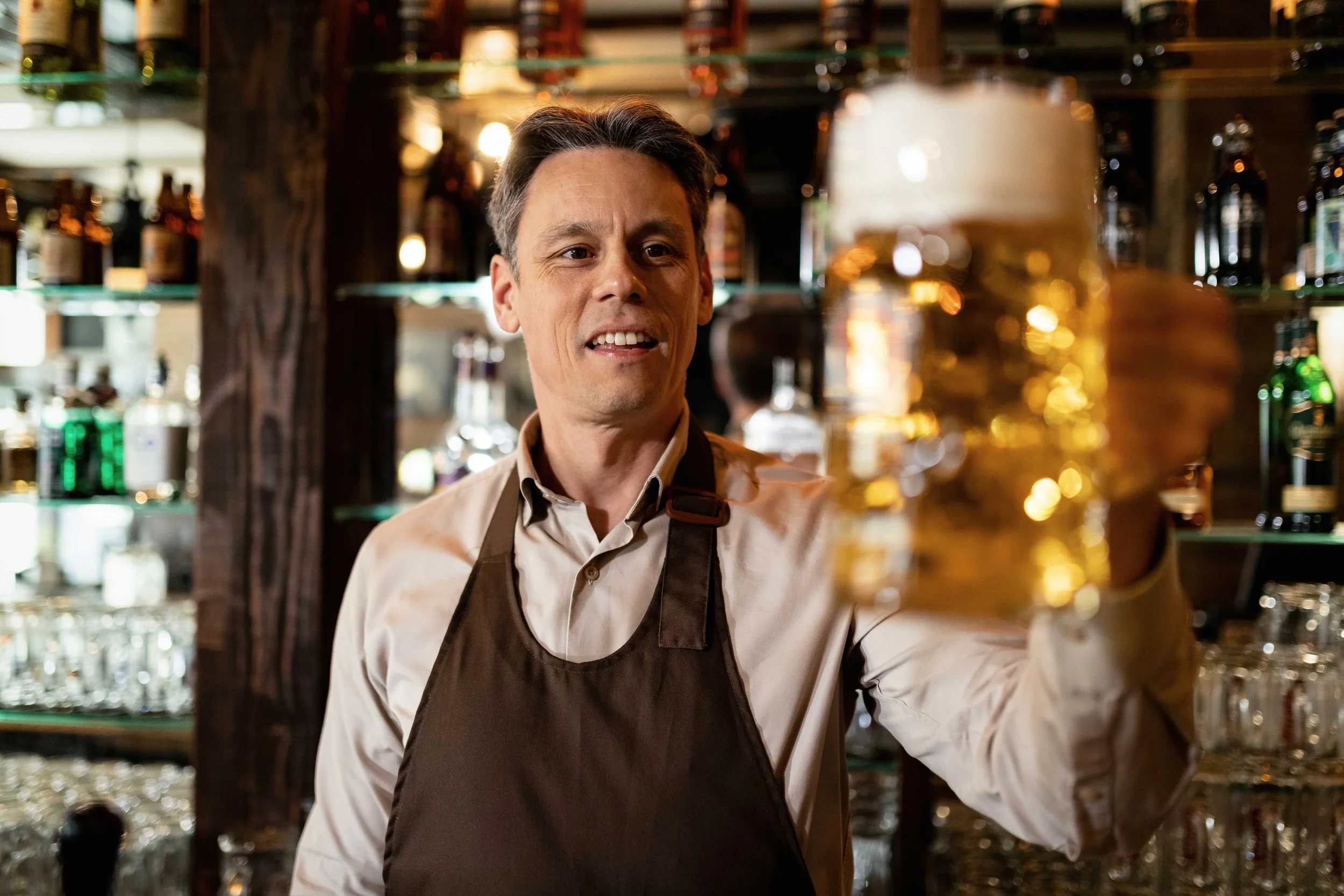 Hombre atendiendo en un bar con anaqueles llenos de botellas y cristalería de fondo.