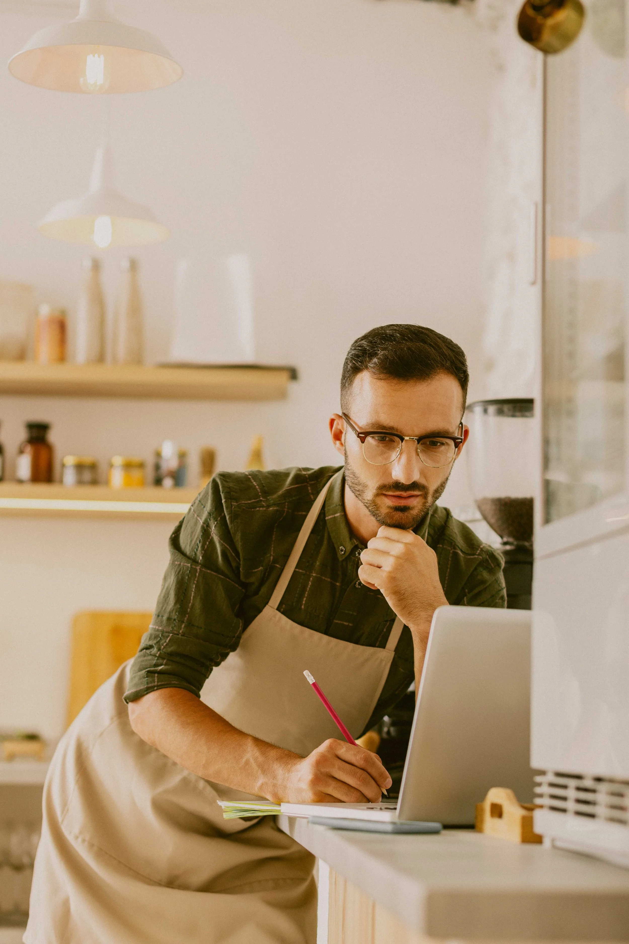Hombre con gafas y barba, usando delantal, revisando una computadora portátil en una cafetería o panadería.