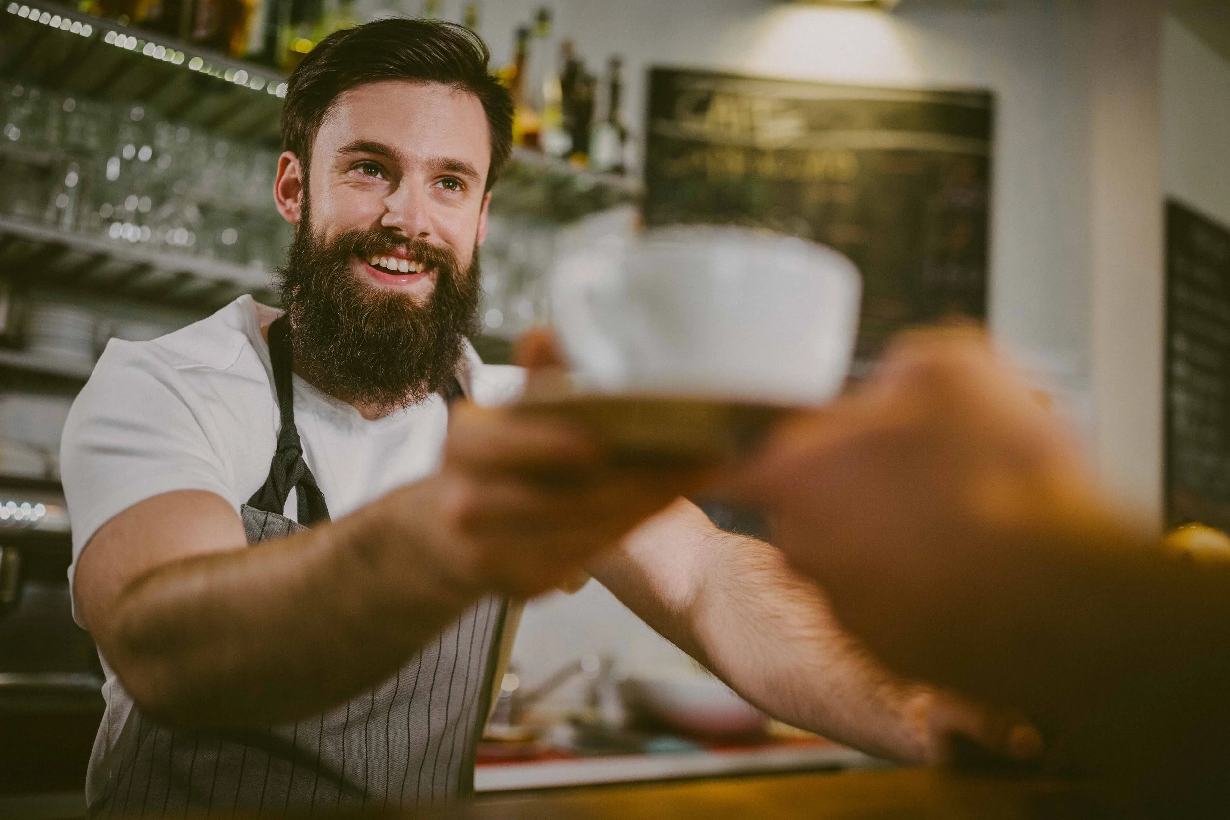 Hombre con barba sonriendo mientras entrega una taza en un ambiente de cafetería