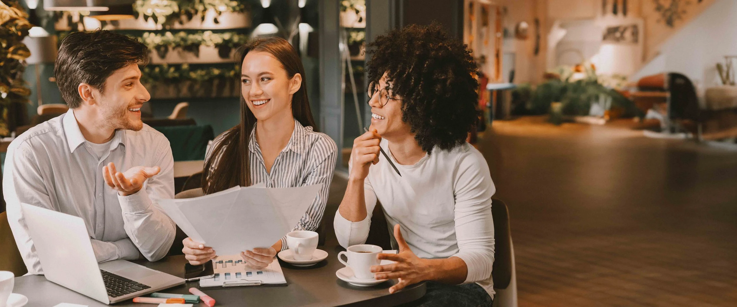 Tres personas en una cafetería discutiendo y sonriendo, con una laptop y papeles sobre la mesa.