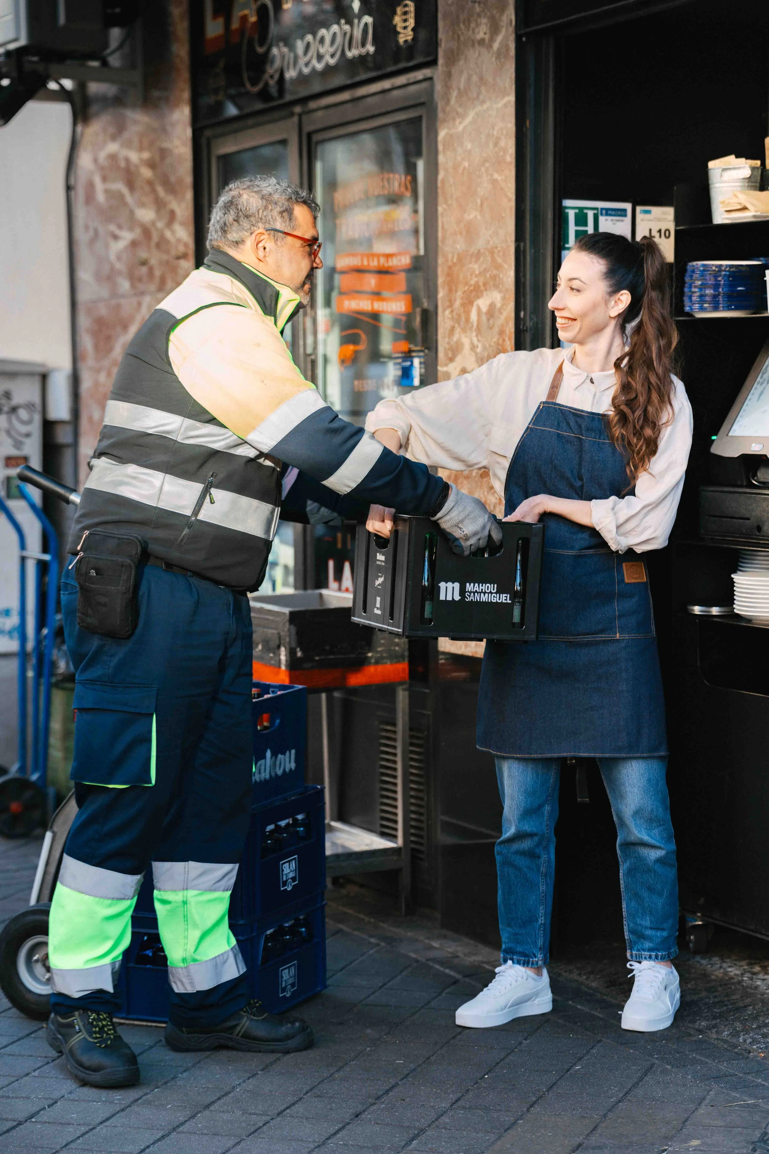 un repartidor entregando un pedido a una empleada de un restaurante, en la calle, con paredes de piedra y carteles en la ventana del negocio.