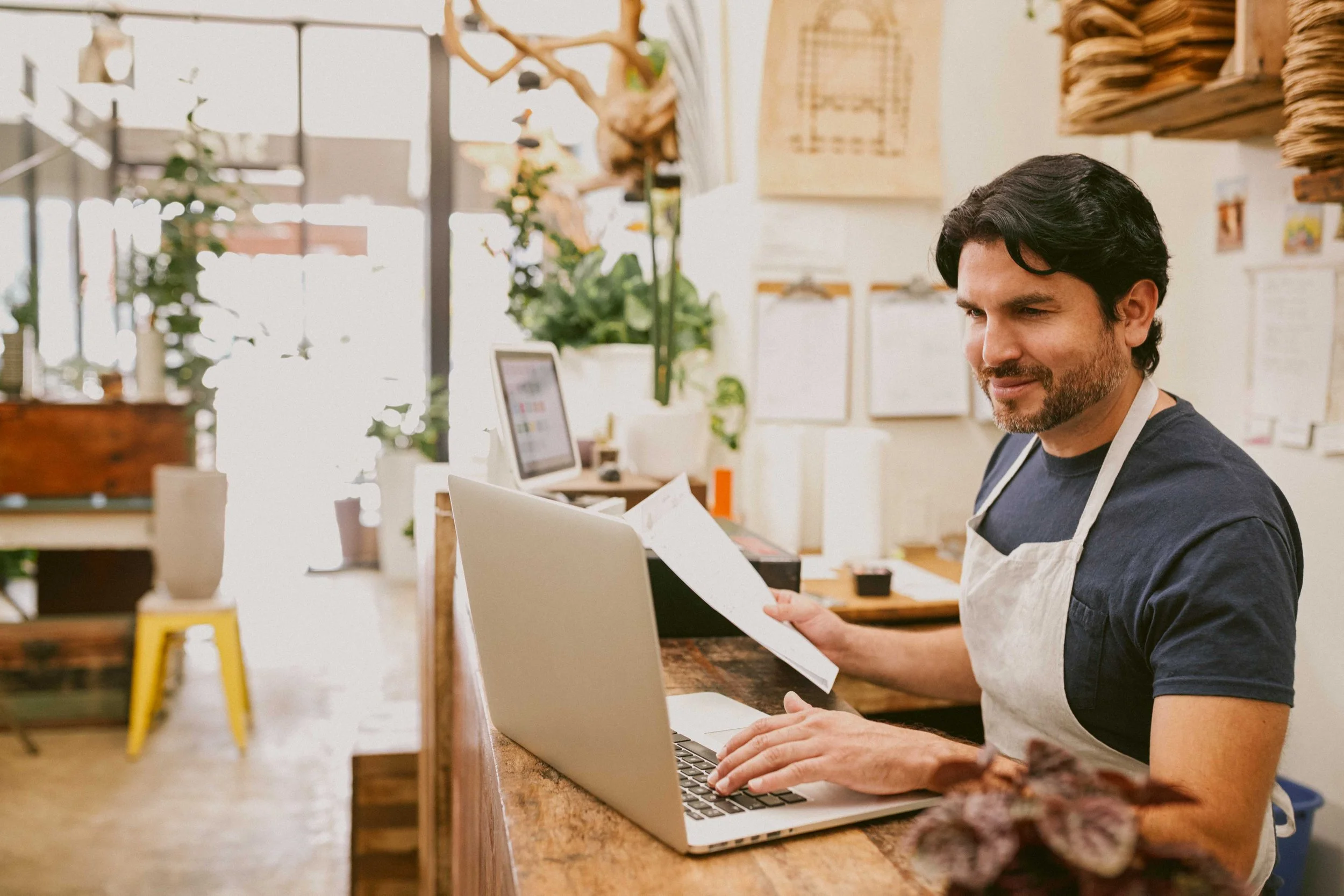 Hombre en una cafetería revisando un recibo o lista, con una computadora portátil abierta frente a él.