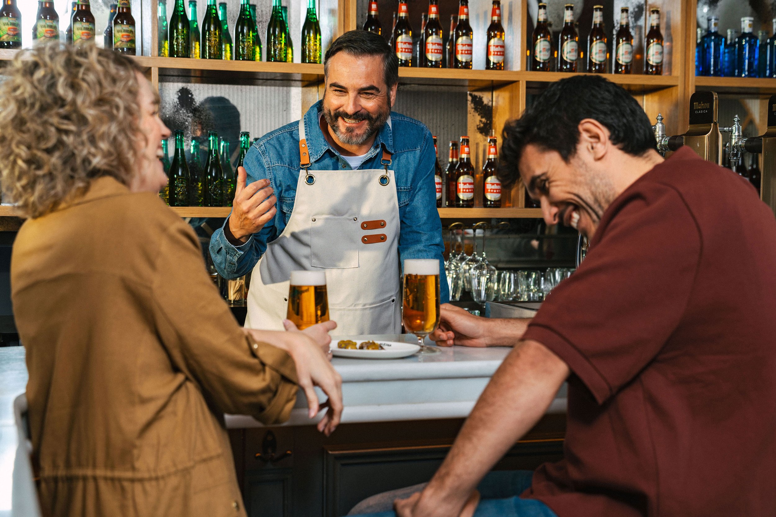 Tres personas conversando en un bar, una mujer y un hombre tomando cerveza y el camarero sonriendo, en el fondo hay estantes con botellas de diferentes colores.