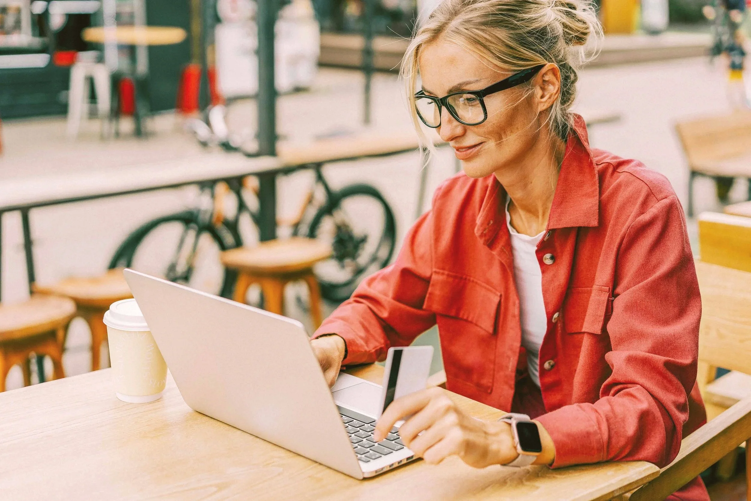Mujer con gafas, camisa roja, usando una laptop y una tarjeta de crédito en una cafetería