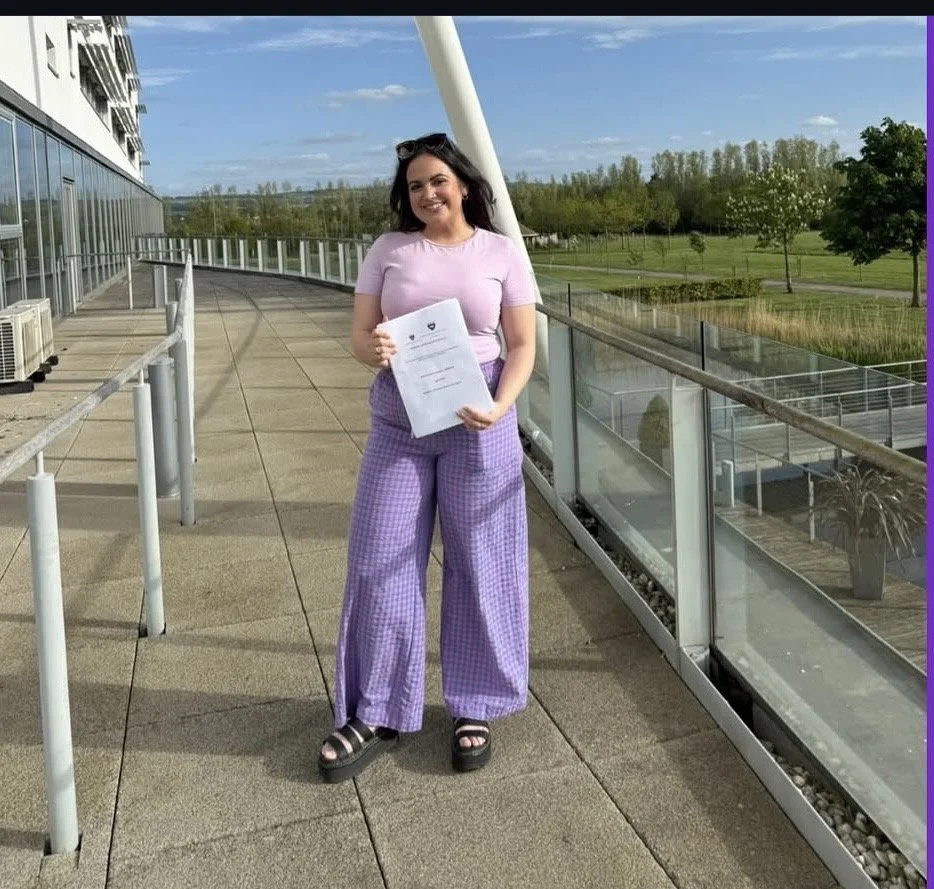 A young woman standing on an outdoor balcony holding a sheet of paper, smiling. She is wearing a light purple t-shirt, purple checkered wide-leg pants, and black platform sandals. The background features a glass building, a railing, and a green park with trees under a partly cloudy sky.