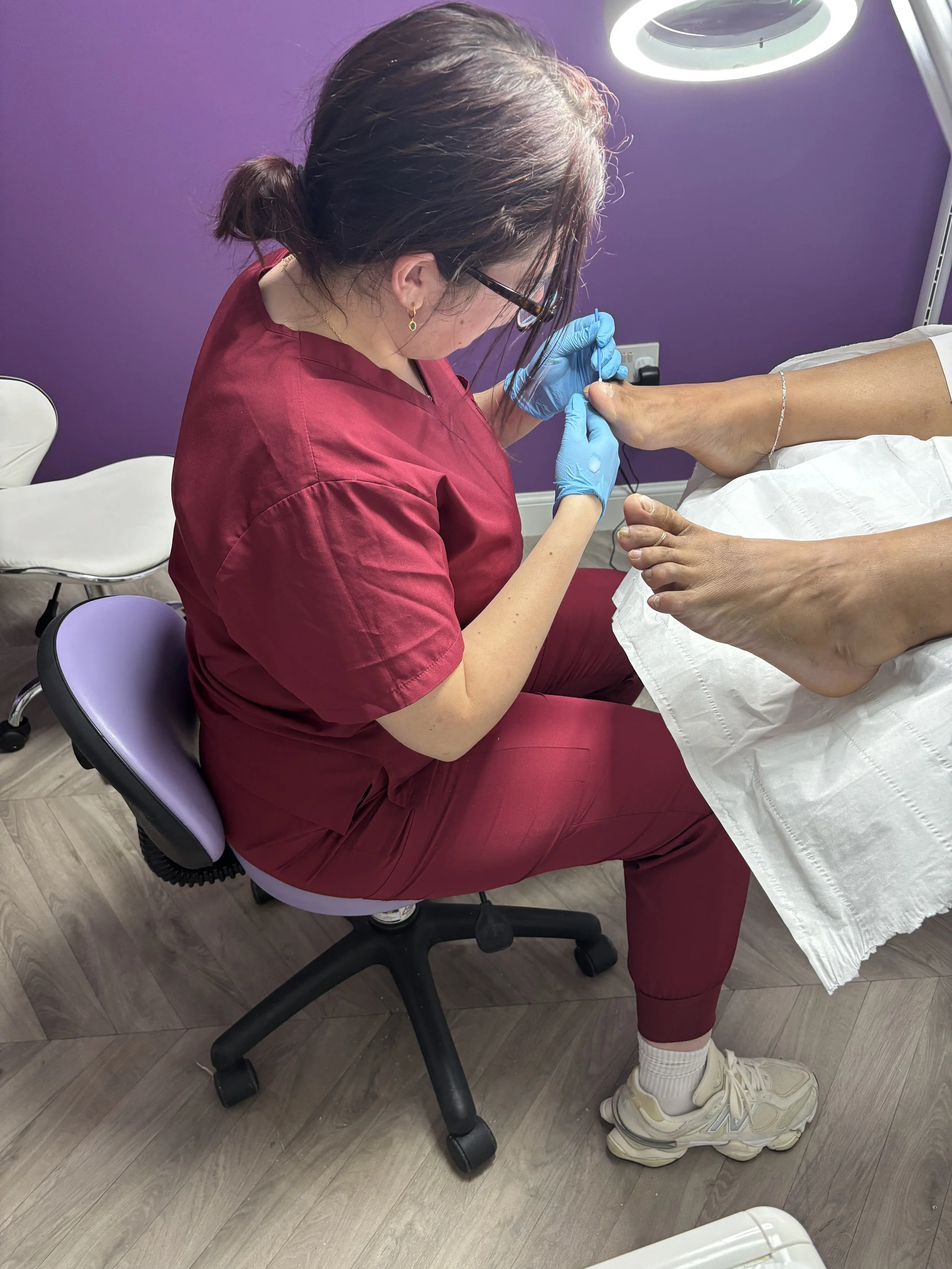 A person receiving a pedicure from a nail technician in a salon with purple walls. The technician, wearing glasses, a red uniform, and blue gloves, is working on the person's toenails. The person is sitting on a black stool, wearing white sneakers, socks, and white shorts. There are chairs and a heater in the background.