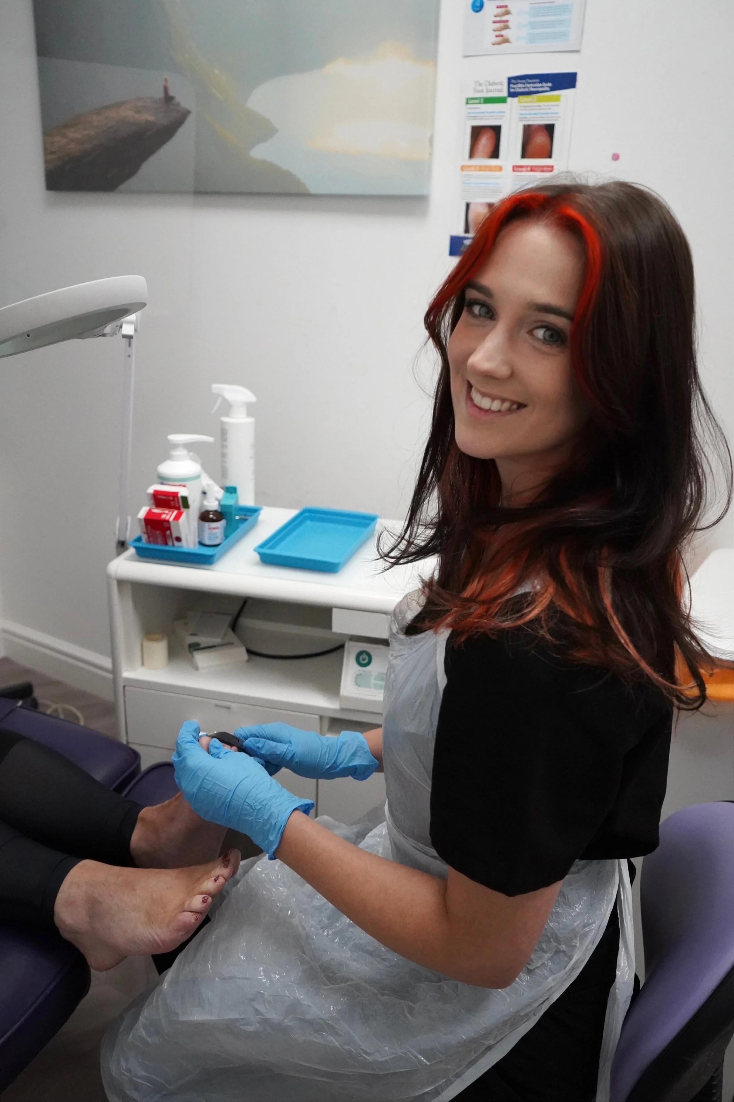A woman with red and black hair in a medical clinic, wearing gloves and assisting a patient with their foot. Medical supplies and informational posters are visible in the background.