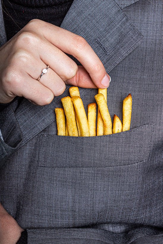 Hand met ring pakt frietjes uit zak van grijs colbert – conceptuele food fotografie mode
