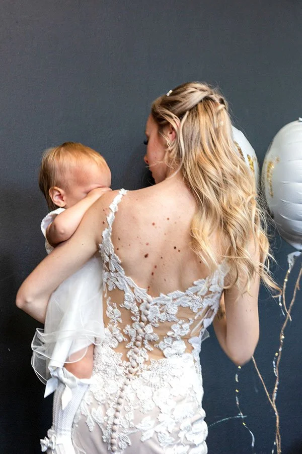 Vrouw in kanten trouwjurk met kind tegen donkere muur en ballonnen – bruidsfotografie met kind