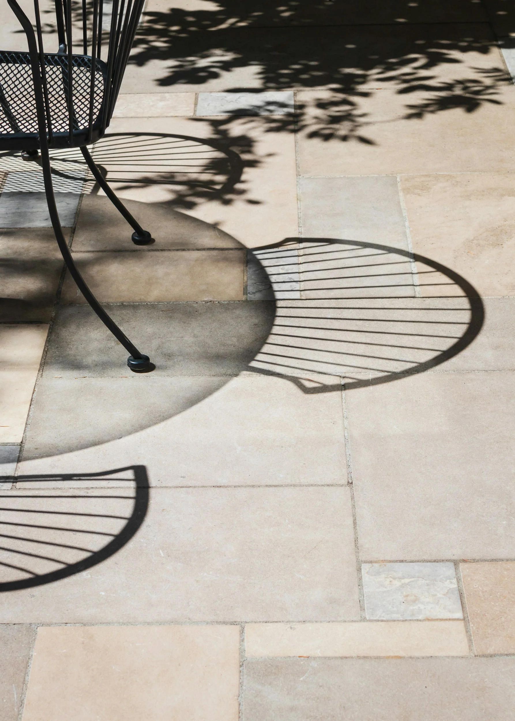 Shadows of metal chairs cast on a tiled outdoor ground with sunlight filtering through leaves.